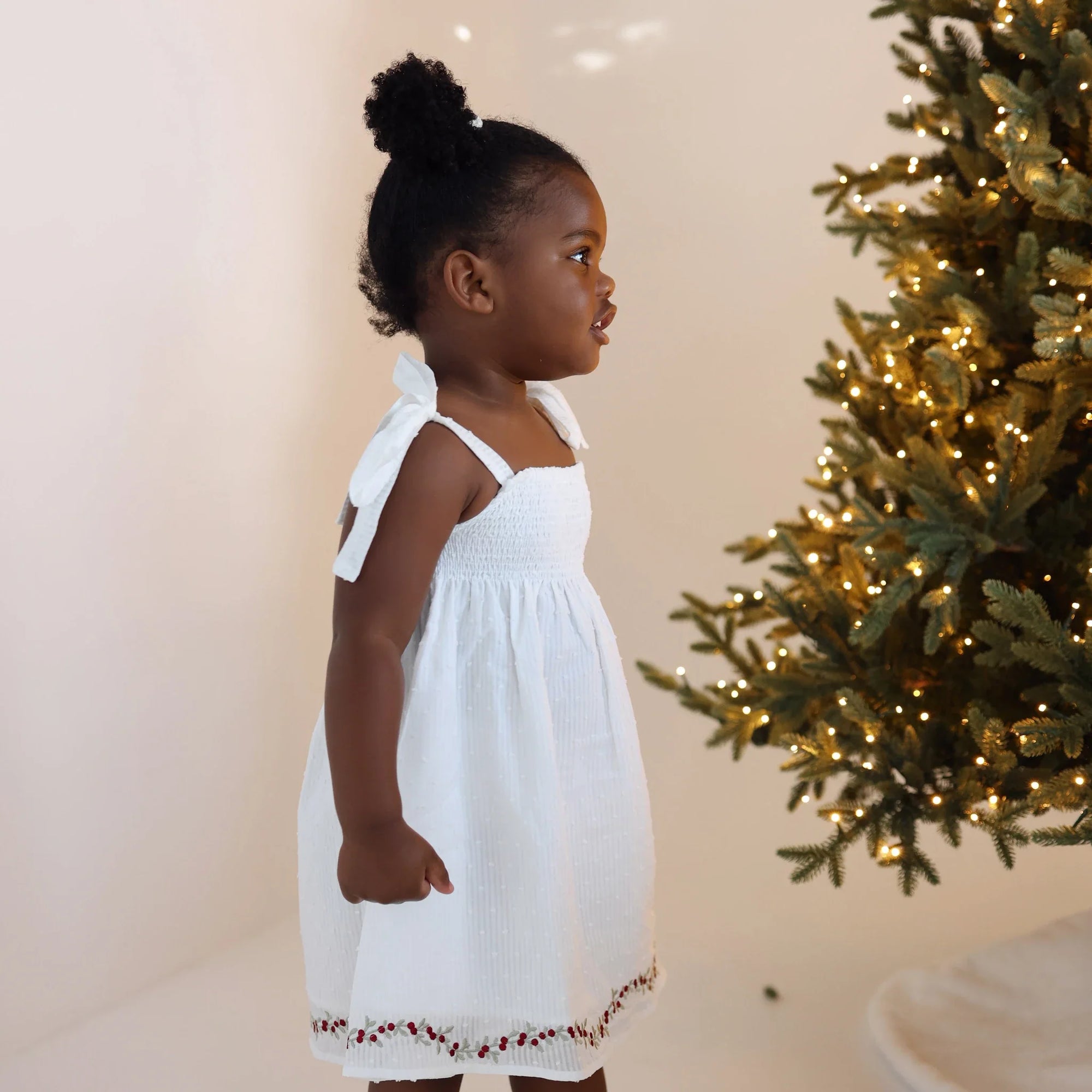 Side profile of a girl in a white shirred Christmas dress with embroidered holly hem beside a lit tree