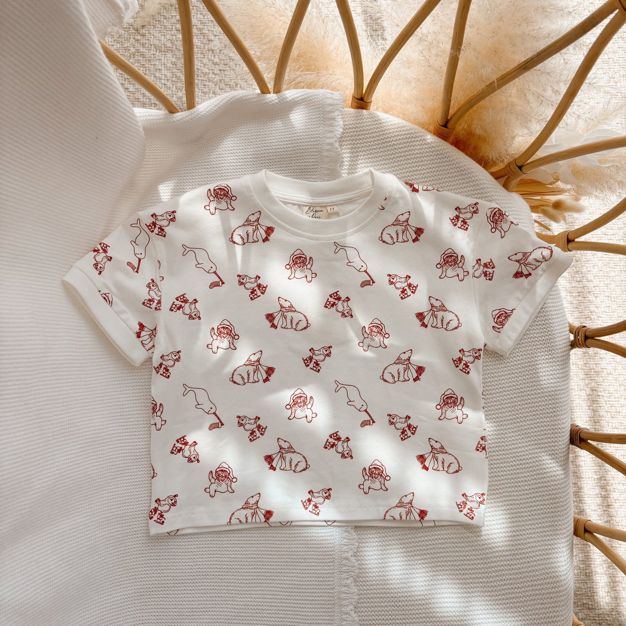 Cream kids tee with an all over red Santa and Christmas character print, flat lay in a bassinet