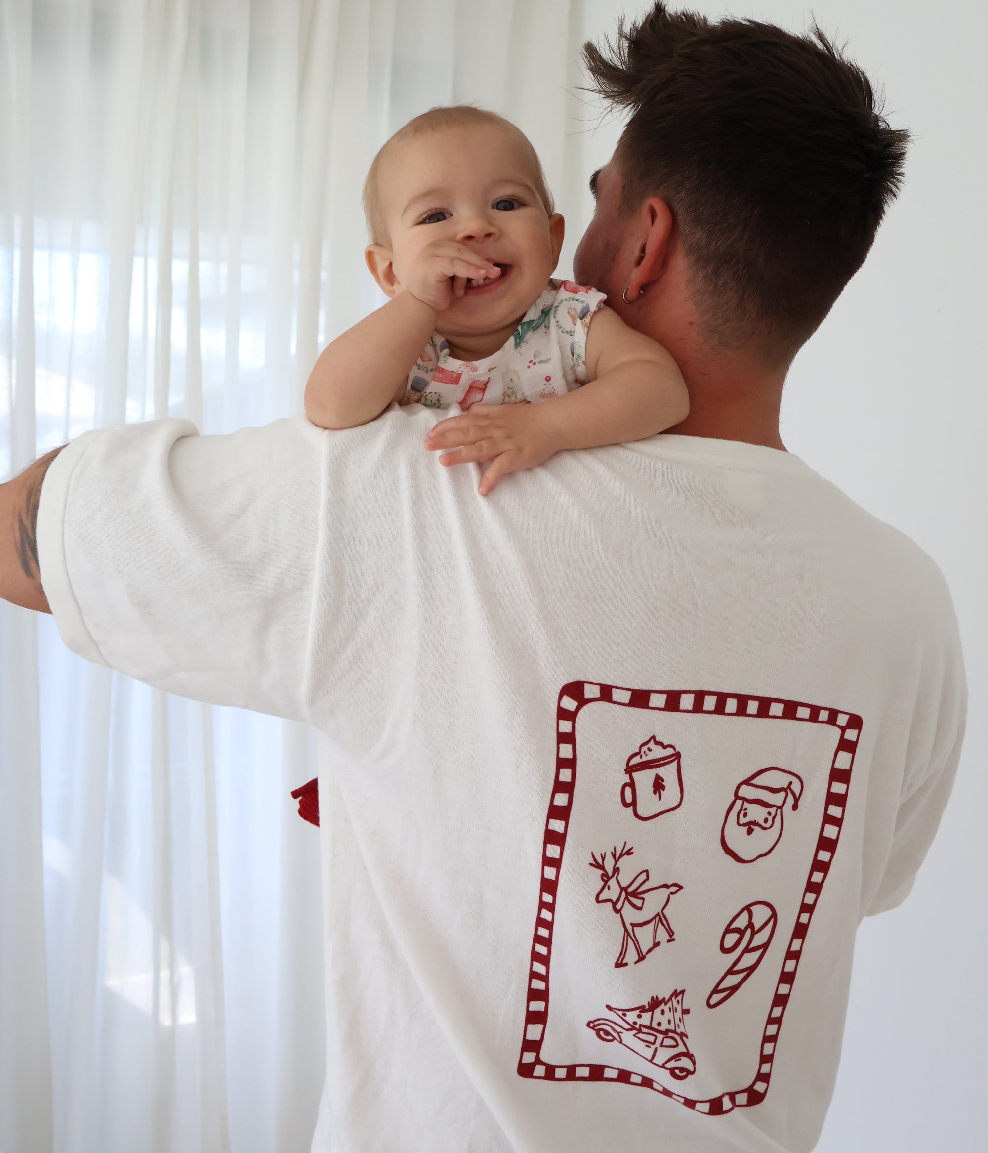 Dad holding a baby over his shoulder, wearing a white tee with a red Christmas doodle back print