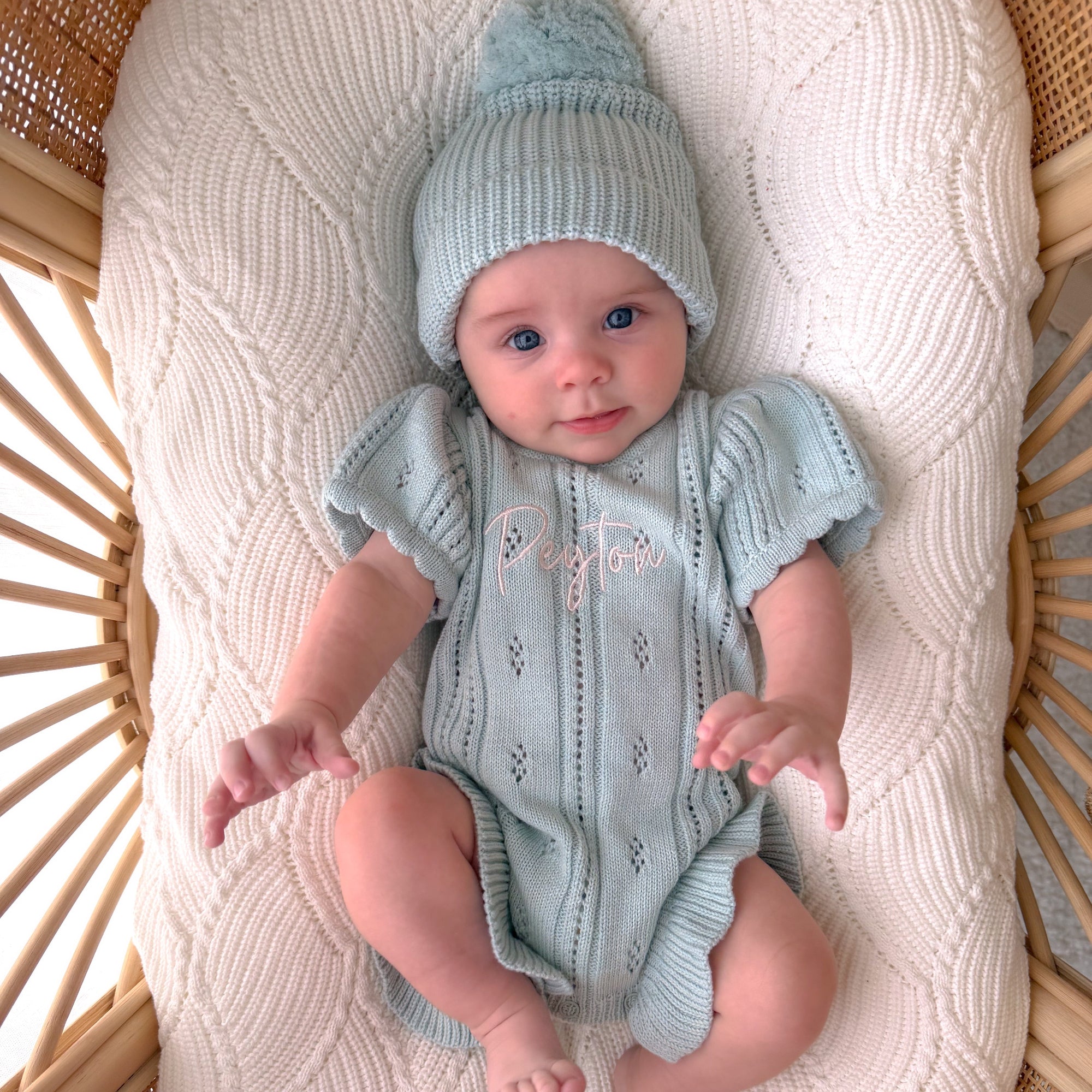 A baby wearing a light blue knit short-sleeve romper and matching beanie hat, lying on a cream cable-knit bassinet cushion inside a wicker bassinet.