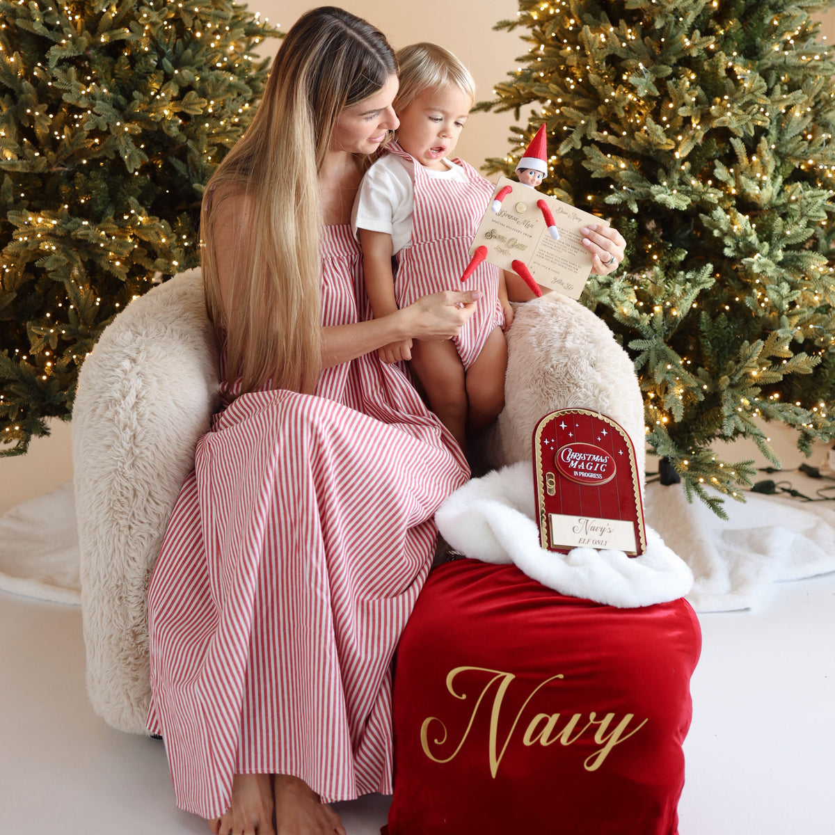 A woman and child in matching red striped outfits sit on a white armchair between two decorated Christmas trees, with a red Santa sack labeled 'Navy' in the foreground.