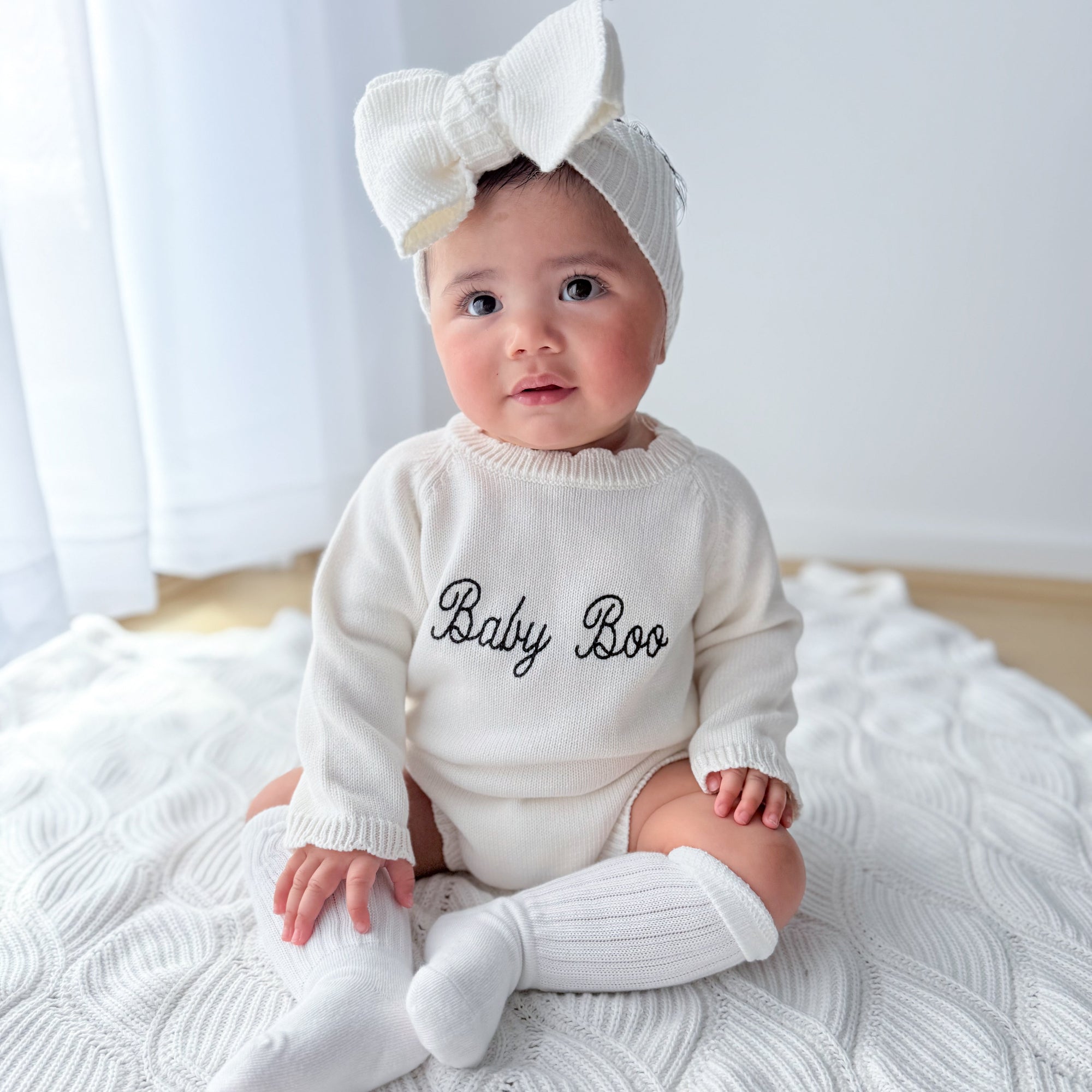 Baby in a cream Baby Boo embroidered knit romper and bow headband sitting on a textured white blanket