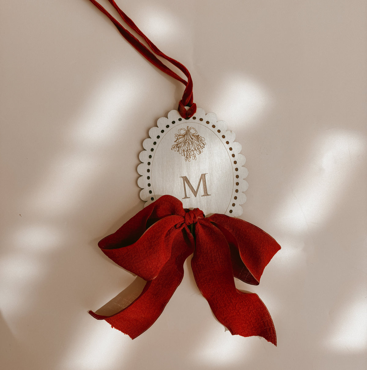 A decorative hanging ornament with a scalloped white oval tag featuring a gold monogram letter 'M' and a gold floral motif, tied with a red ribbon and finished with a large red fabric bow. The ornament is photographed against a warm beige background with soft dappled light shadows.