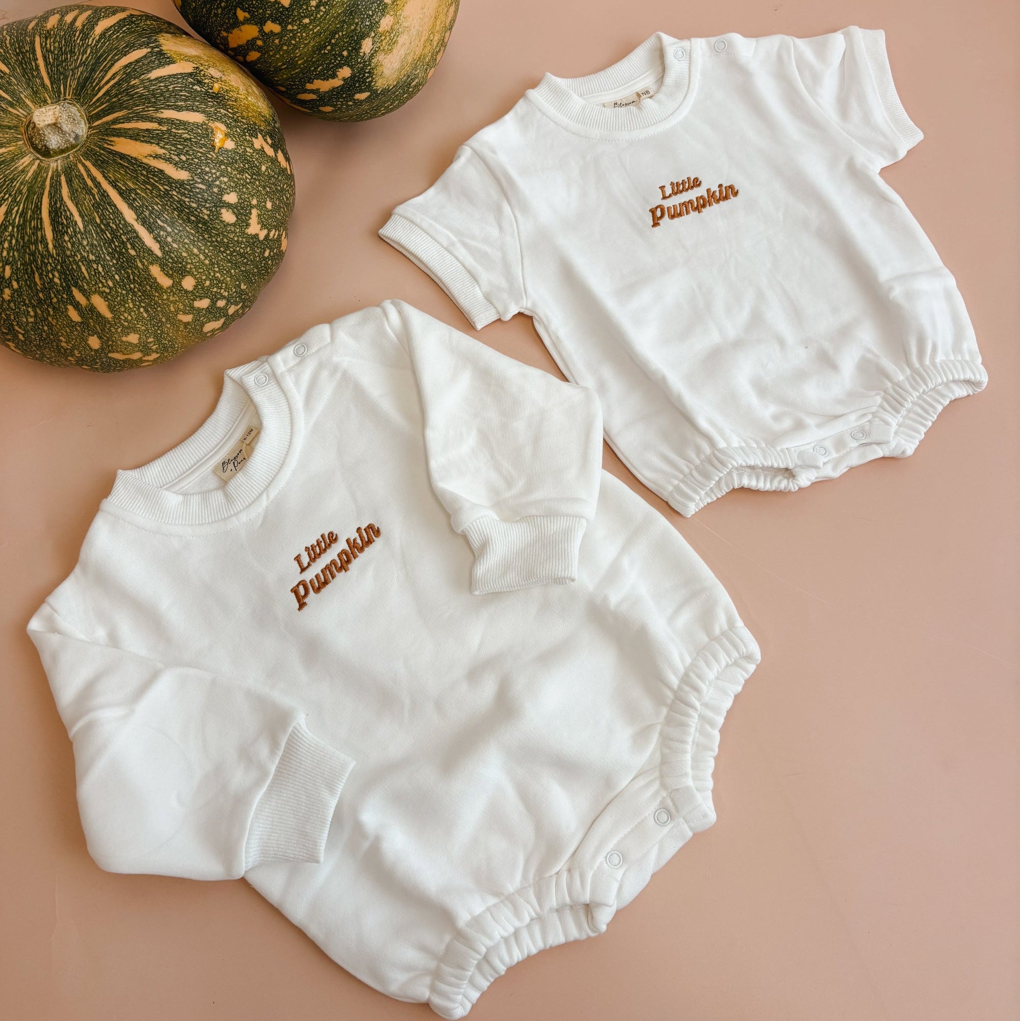 Two white baby bodysuits with 'Little Pumpkin' embroidered in brown on the chest, arranged on a dusty pink background next to two green pumpkins.