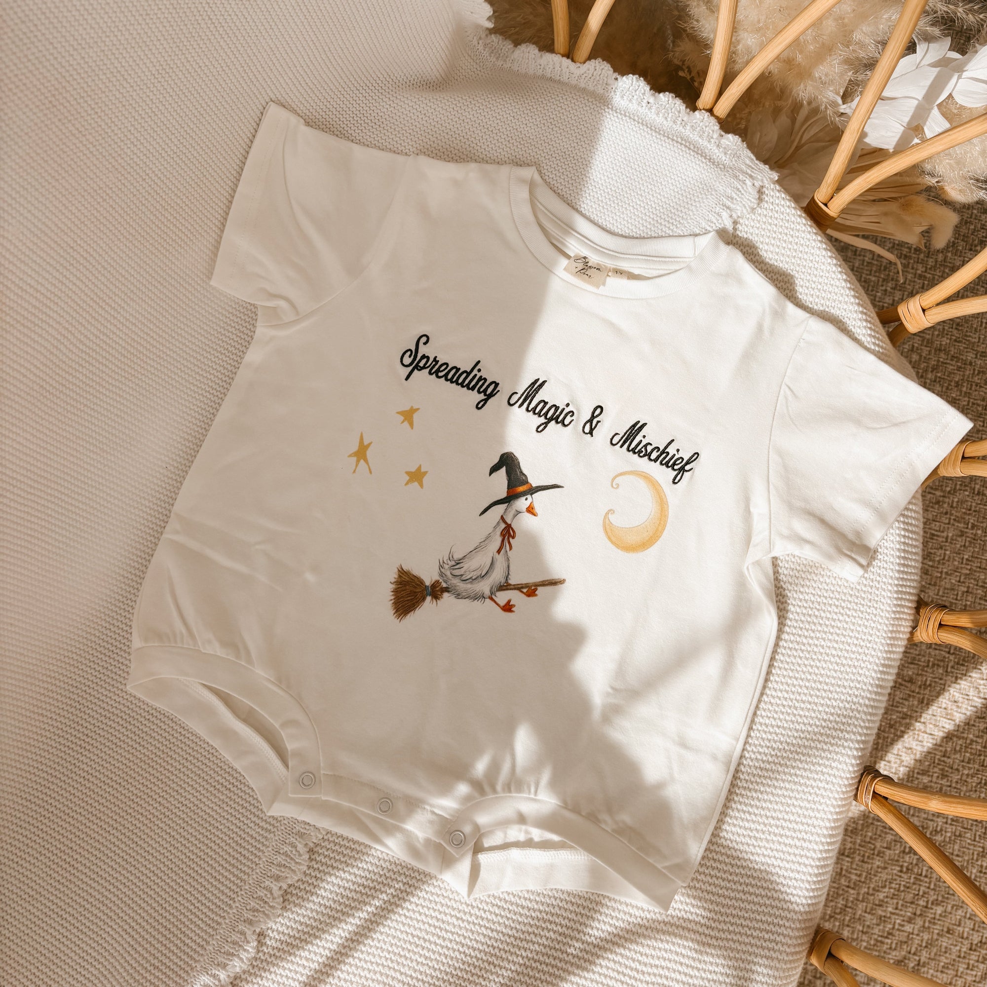 A white short-sleeve baby bodysuit laid flat on a textured cream blanket, featuring a graphic of a witch riding a broomstick under a crescent moon and stars with the text 'Spreading Magic &amp; Mistakes'.
