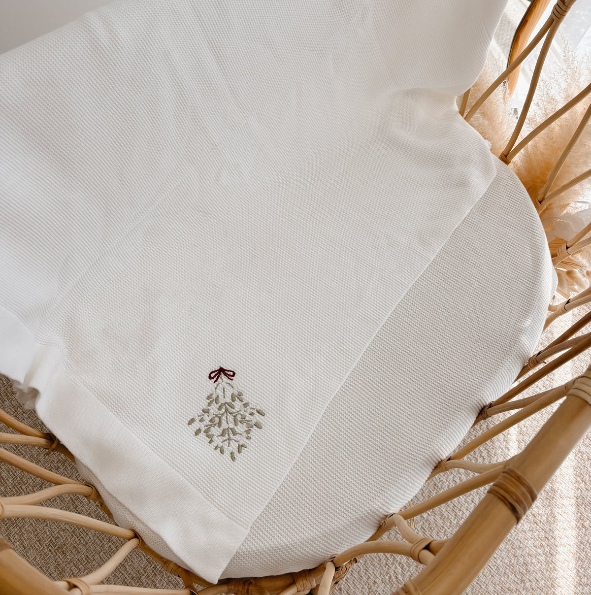 White knit baby blanket embroidered with mistletoe and a red bow, draped over a rattan bassinet