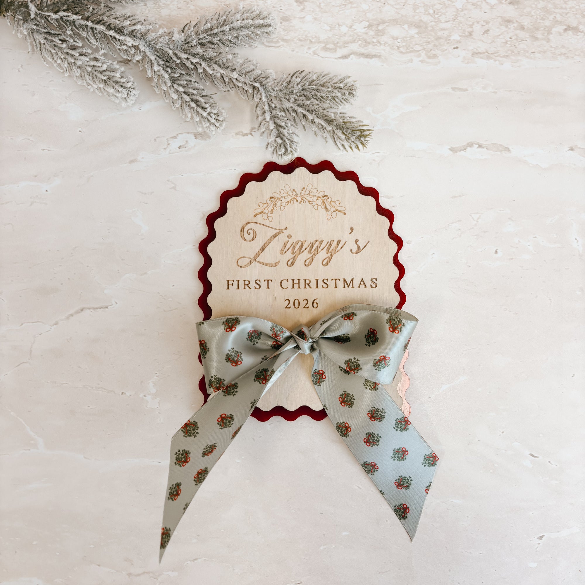 A personalized Christmas ornament tag with a scalloped red border and gold script reading 'Ziggy's First Christmas 2026,' tied with a patterned ribbon, photographed on a light stone surface with a frosted pine branch in the corner.