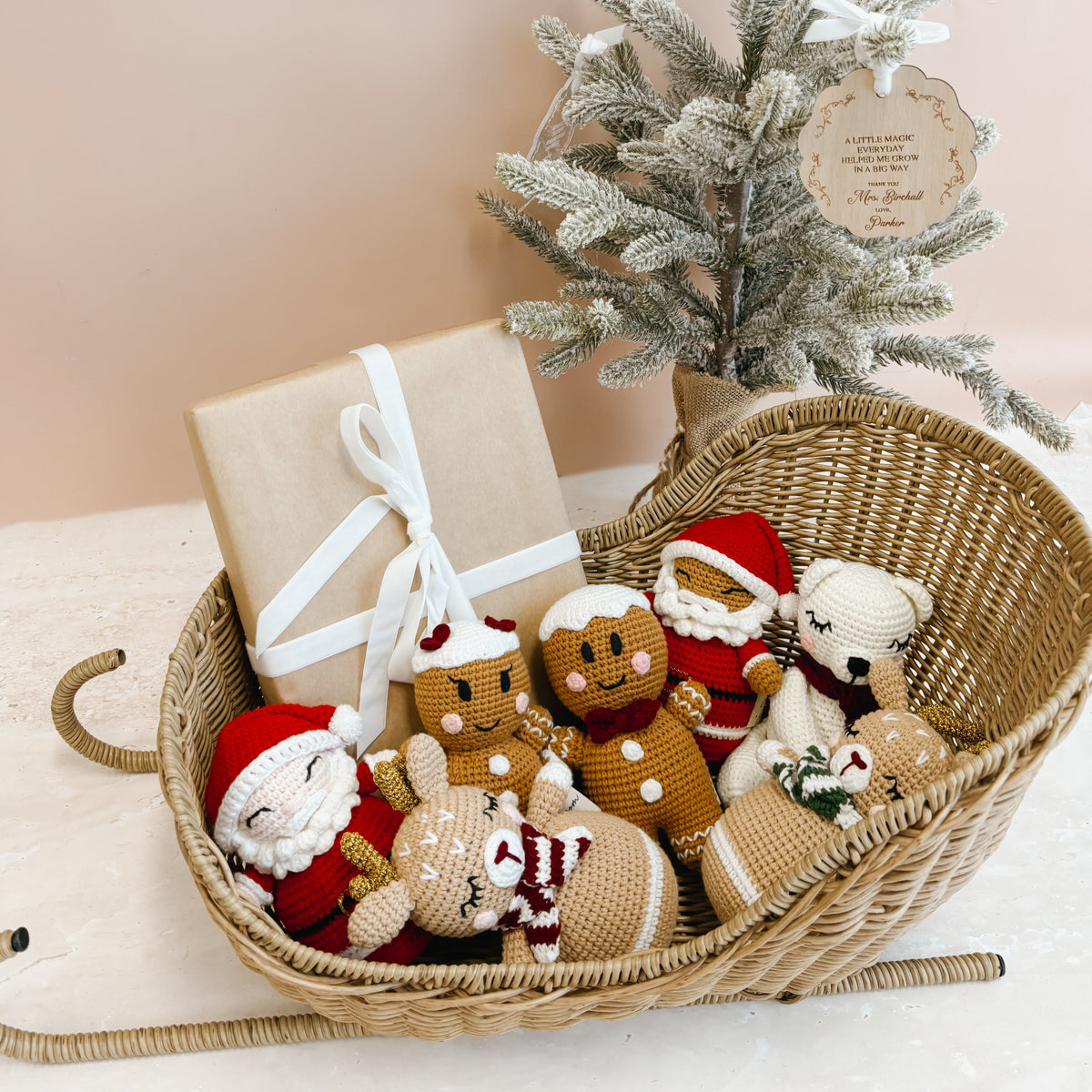 A wicker sled filled with handmade Christmas toys and a kraft gift box tied with a white ribbon, styled beside a small frosted tree against a soft blush background.