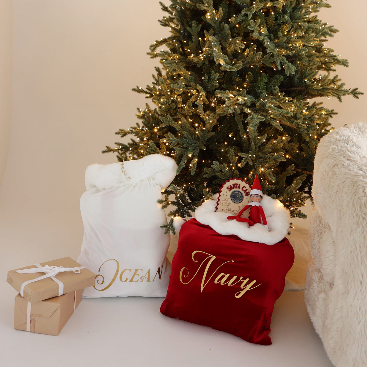 Personalised ivory velvet Santa sack with the name Ocean and burgundy velvet Santa sack with the name Navy under a Christmas tree with an elf
