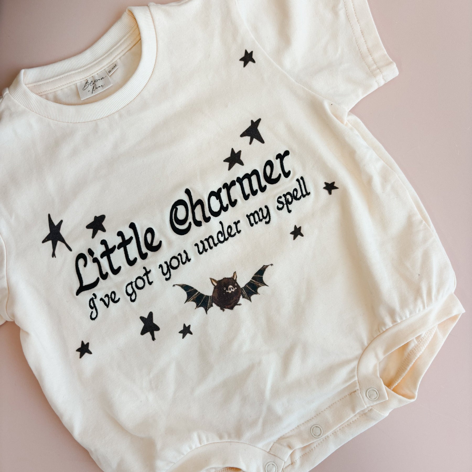 An off-white short-sleeve baby bodysuit with black text reading 'Little Charmer I've got you under my spell' and a small bat graphic, laid flat on a dusty pink background.