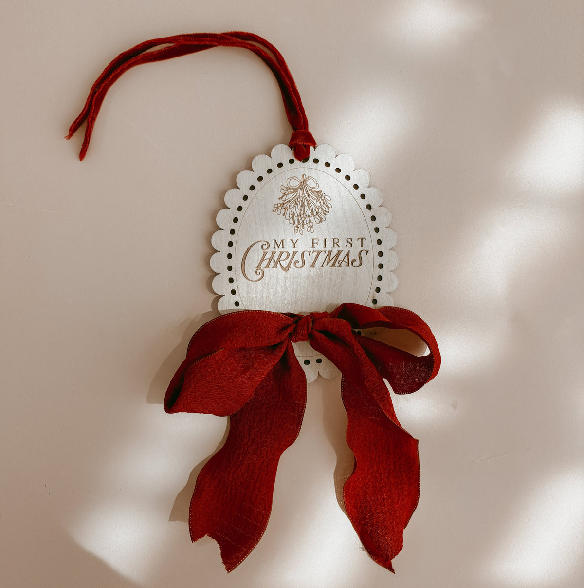 A white scalloped oval Christmas ornament with the words 'MY FIRST CHRISTMAS' and a small tree illustration, hanging from a red ribbon loop with a large red bow tied around the middle, set against a warm beige wall with soft dappled light shadows.