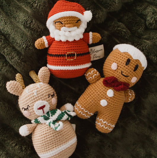 Three handmade crocheted Christmas characters—a Santa Claus, a gingerbread man, and a reindeer—arranged on a dark green textured blanket. A small fabric tag is visible on the Santa's sleeve.