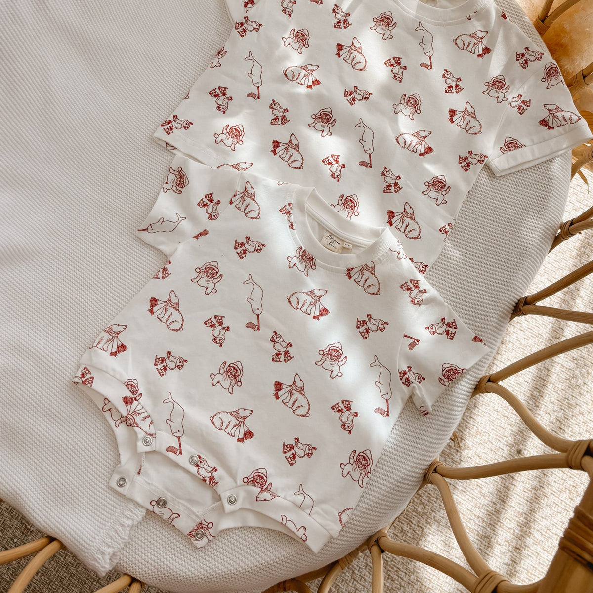 Matching Santa print kids tee and baby romper in cream with red print, flat lay in a bassinet
