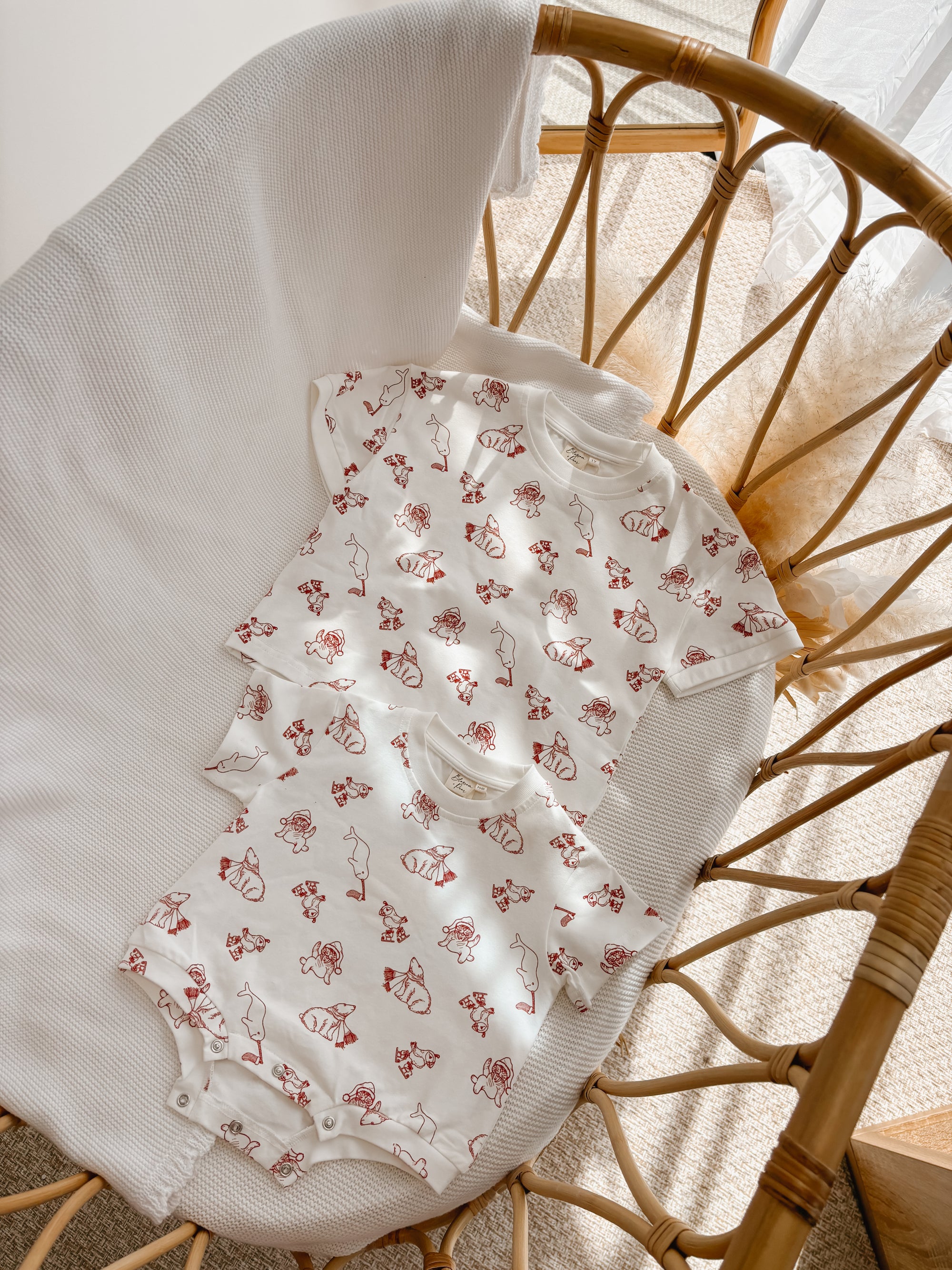 Matching Santa print kids tee and baby romper in cream with red print, flat lay in a bassinet