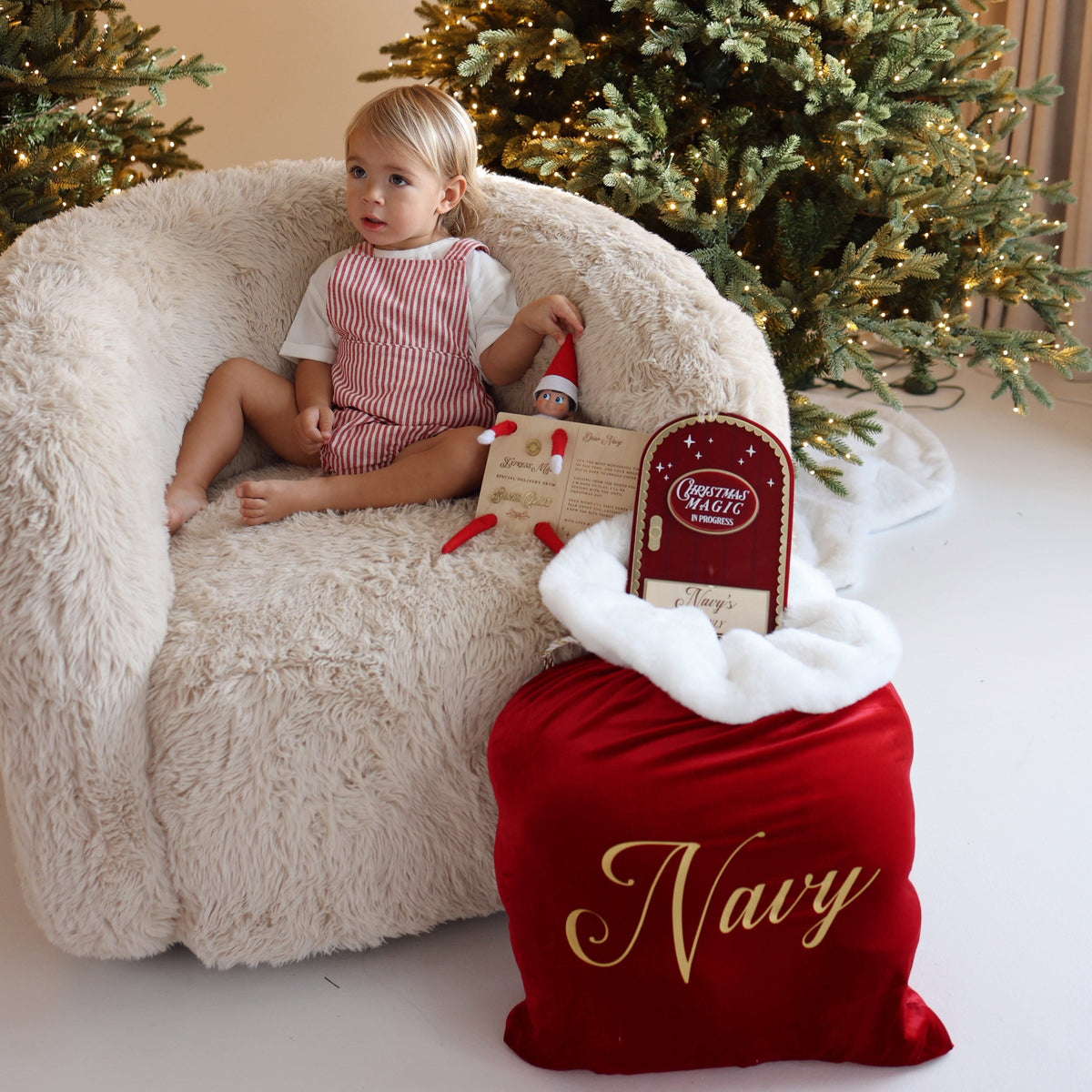 A toddler sits on a fluffy cream armchair in front of decorated Christmas trees, holding a small red-and-white Santa hat. In the foreground is a red Santa sack with white trim and gold script reading 'Navy,' plus a small gift box with a 'Merry Christmas' tag.