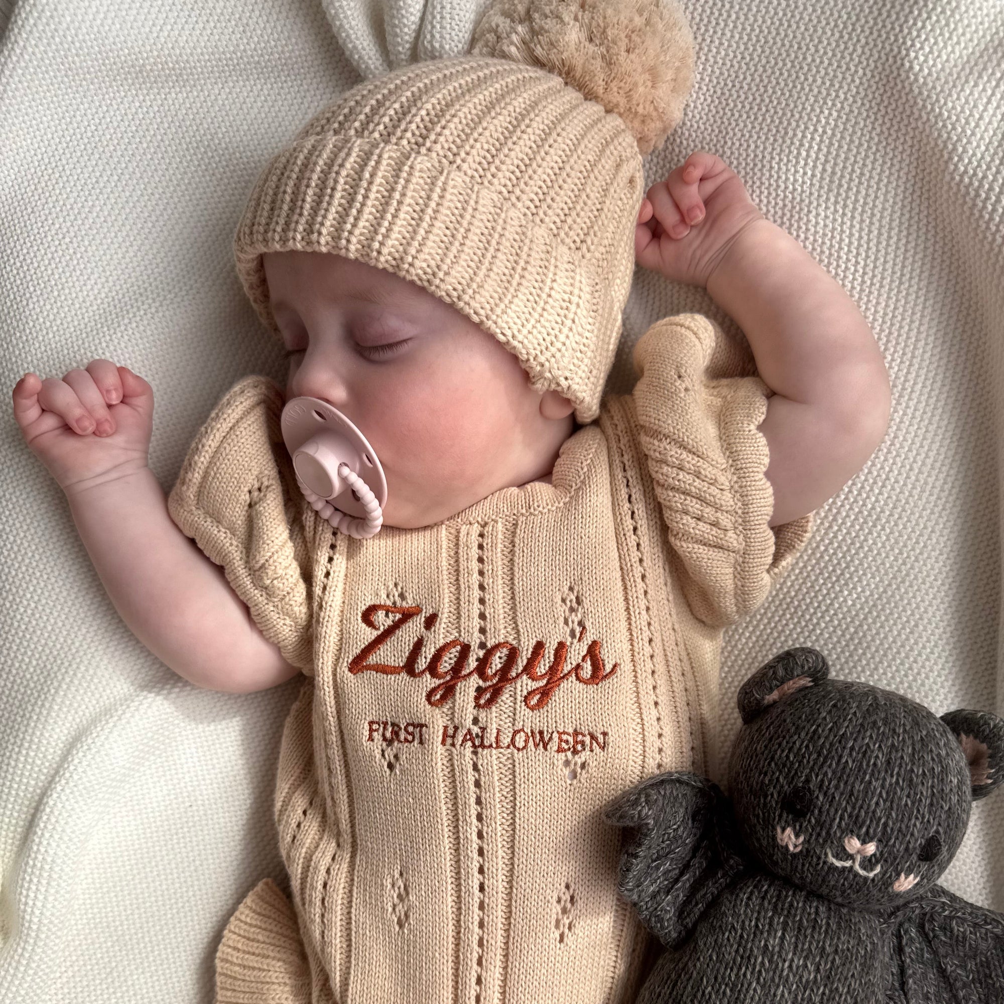 Sleeping baby in a personalised First Halloween knit romper and pom beanie beside a knitted bat toy