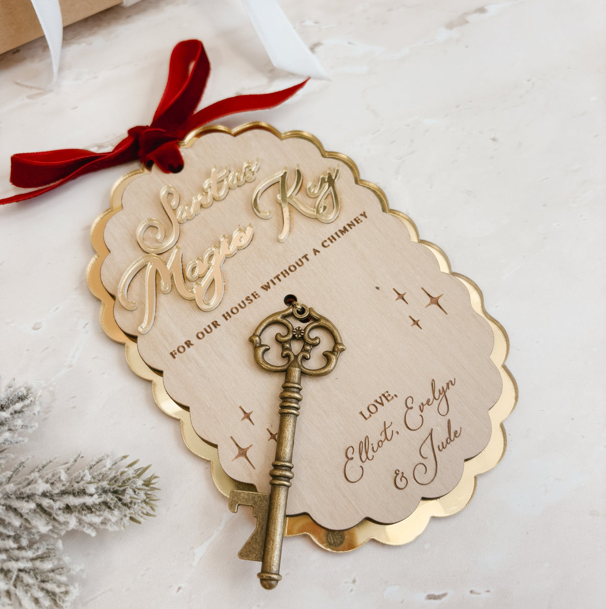 Personalized wooden Santa Magic Key ornament with gold foil lettering, a red ribbon, and a vintage-style key, displayed on a light surface with a wrapped gift and white faux snow in the background.