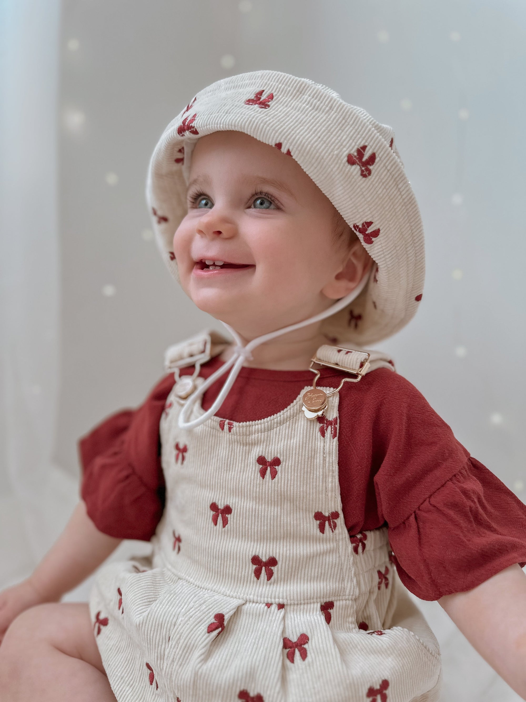 Smiling baby in a red bow print corduroy overall romper and matching bucket hat