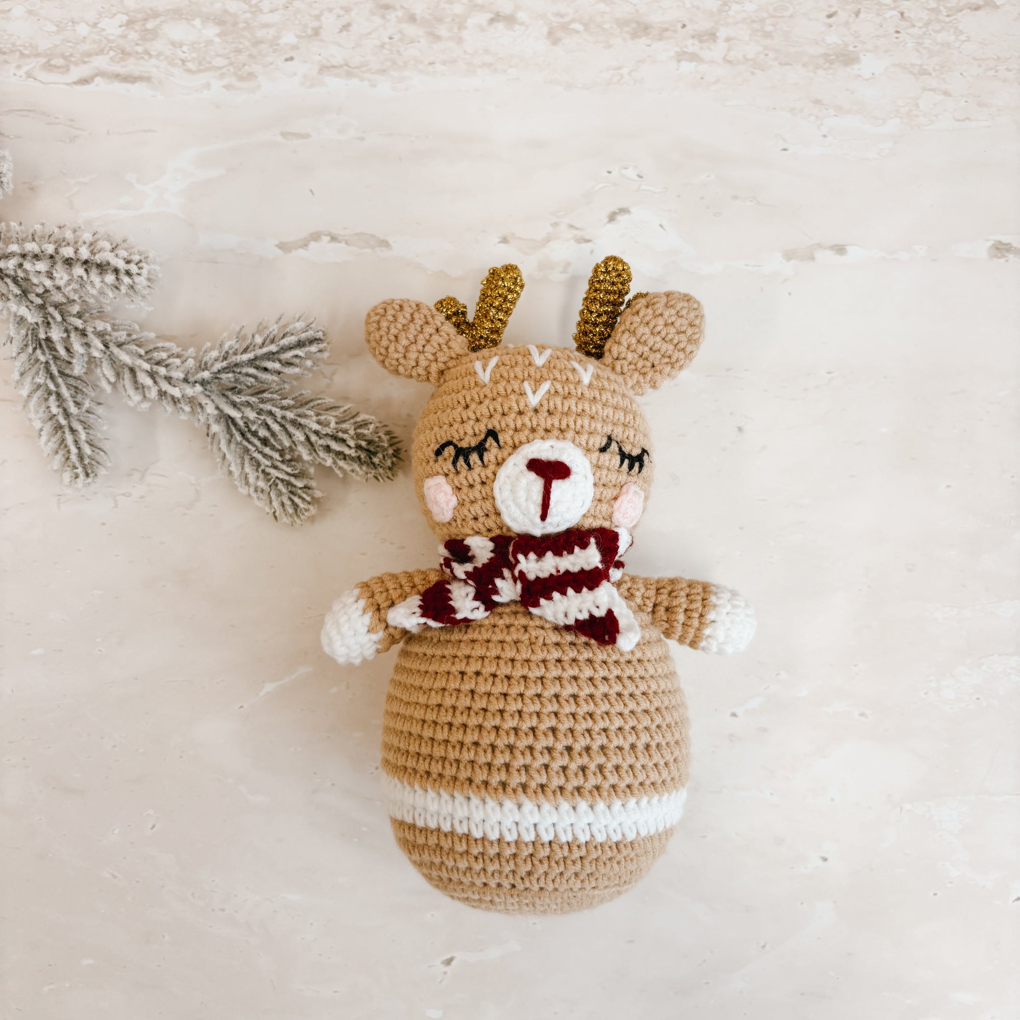 A crocheted plush reindeer toy with gold antlers, a brown and white striped body, and a maroon and white striped scarf, lying on a textured beige surface next to a decorative pine branch.