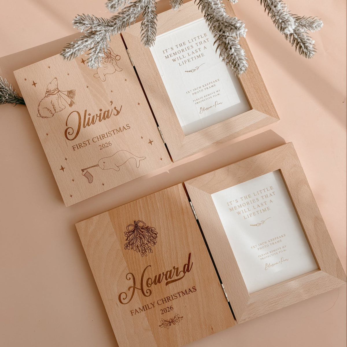 Two open personalised wooden keepsake photo frames laid flat, engraved 'Olivia's First Christmas 2026' with polar bear and seal, and 'Howard Family Christmas 2026' with mistletoe, styled with frosted pine