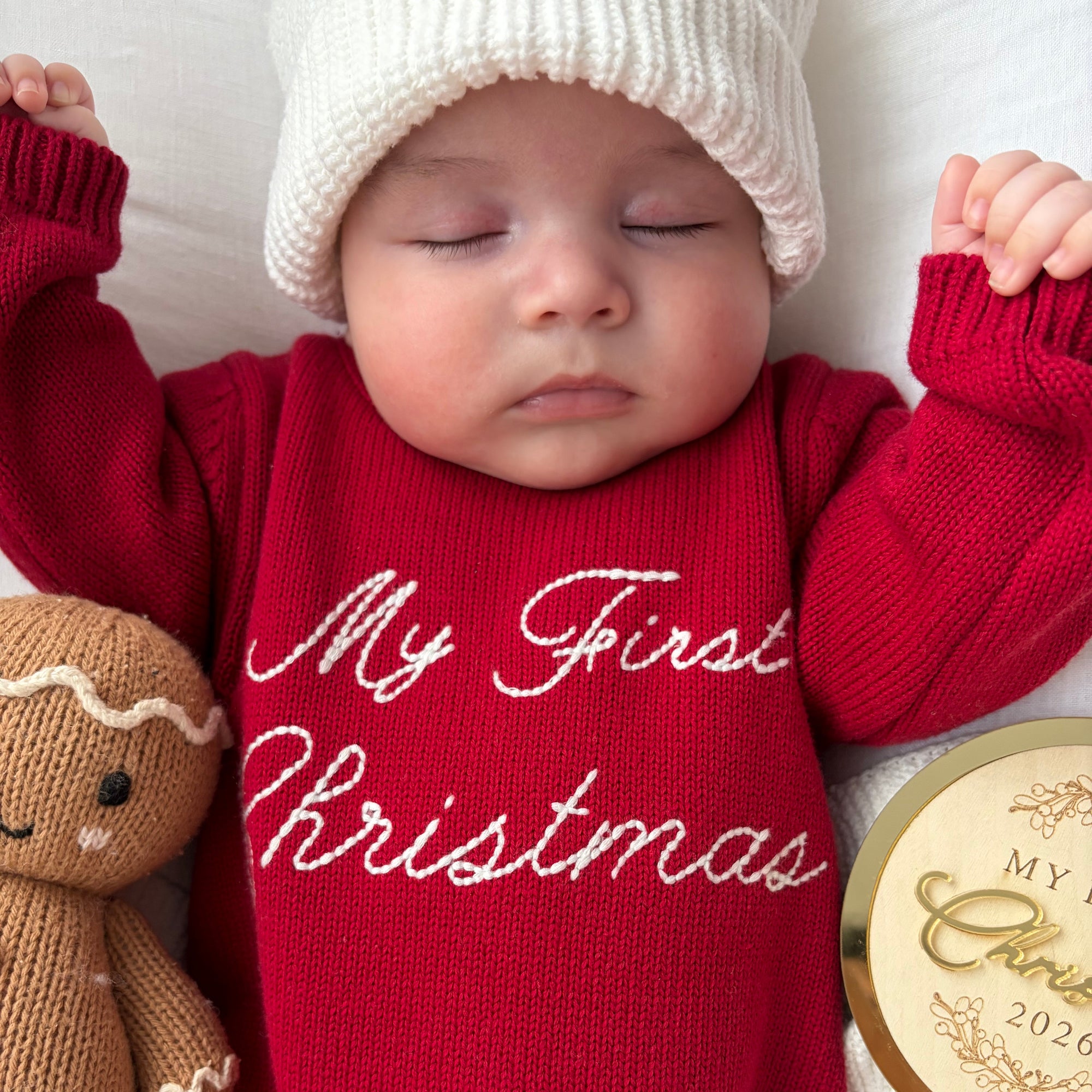 Close up of a sleeping baby in a red knit My First Christmas romper and white beanie beside a gingerbread toy