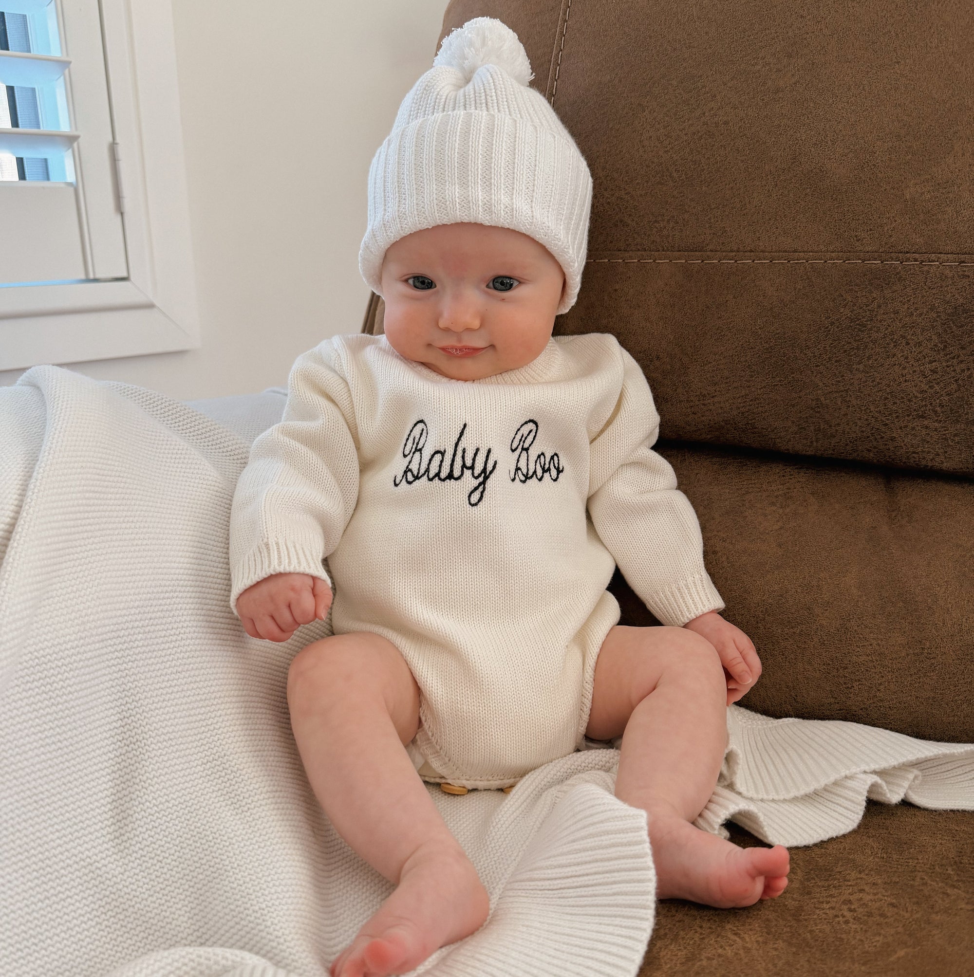 Baby wearing a cream knit romper embroidered with 'Baby Boo' in navy script and a white pom beanie, sitting against a leather lounge