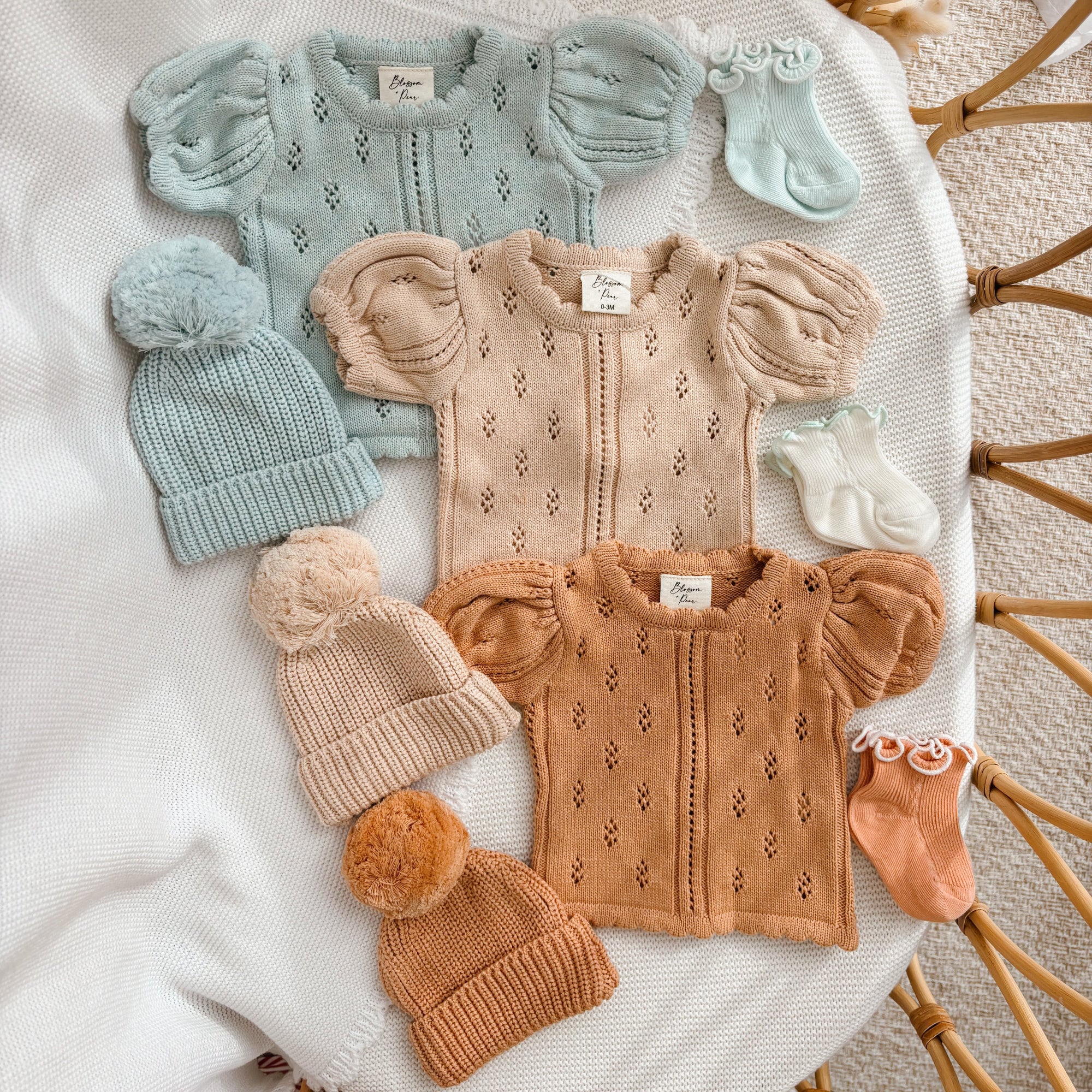 Margot pointelle knit baby rompers in Periwinkle Blue, Vanilla Oat and Pumpkin with pom beanies in a rattan bassinet