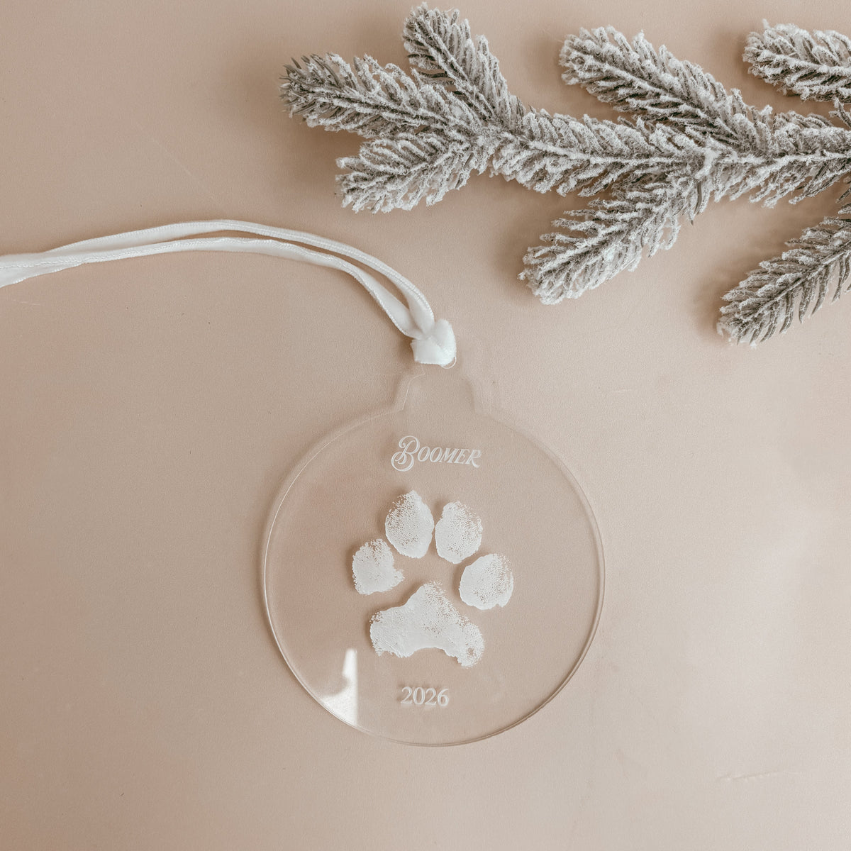 Personalised clear acrylic pet paw print Christmas ornament engraved with the pet's name and 2026, styled with a frosted pine branch