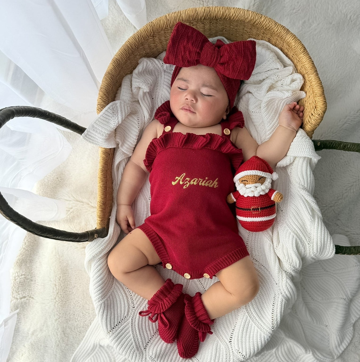 A newborn baby wearing a red knit romper, matching bow headband, and booties, lying in a wicker bassinet with a white blanket. The romper is personalized with the name 'Azariah' in gold embroidery.