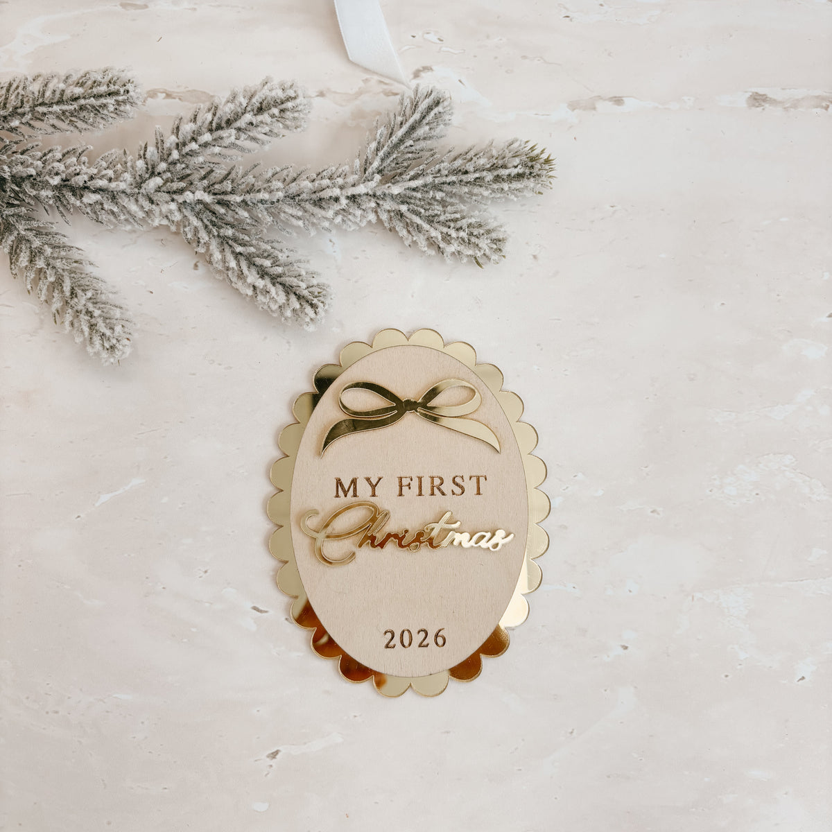 A scalloped oval Christmas ornament tag with the text 'MY FIRST Christmas 2026' in gold foil, styled on a light textured surface with a flocked pine branch and part of a kraft gift box with a white ribbon.