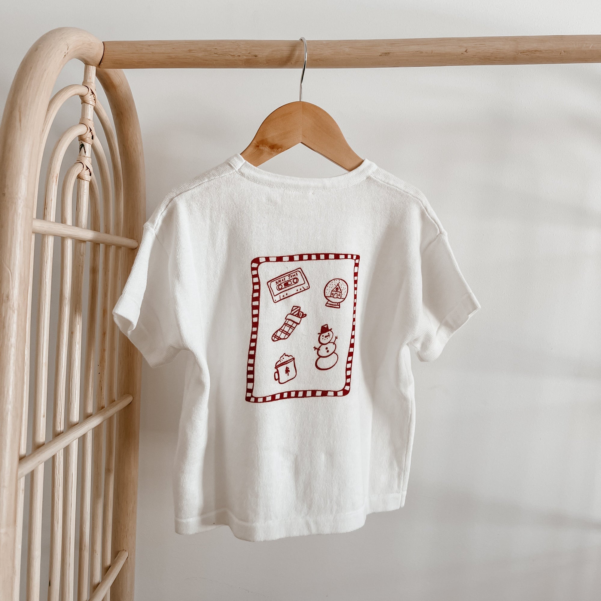 White kids tee with a red Christmas doodle print of a snow globe, stocking and snowman, on a hanger
