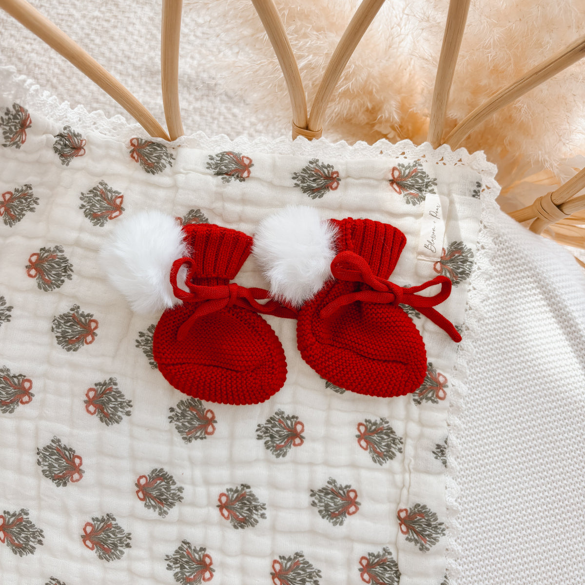 Pair of red knit baby booties with white pom poms on a wreath print muslin blanket