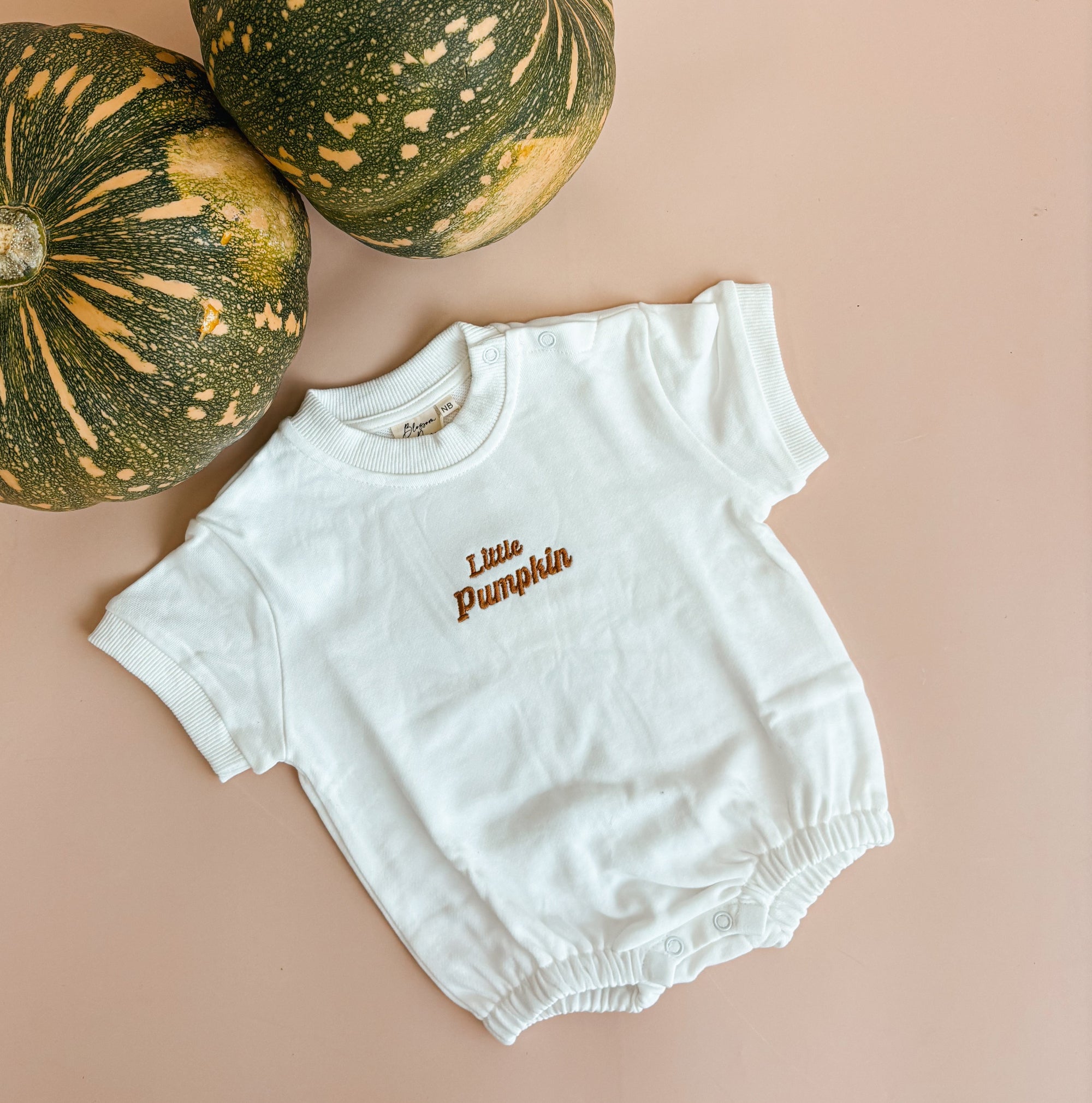 A white short-sleeve baby romper with 'Little Pumpkin' printed in brown on the chest, styled on a dusty pink background next to two green and yellow striped pumpkins.
