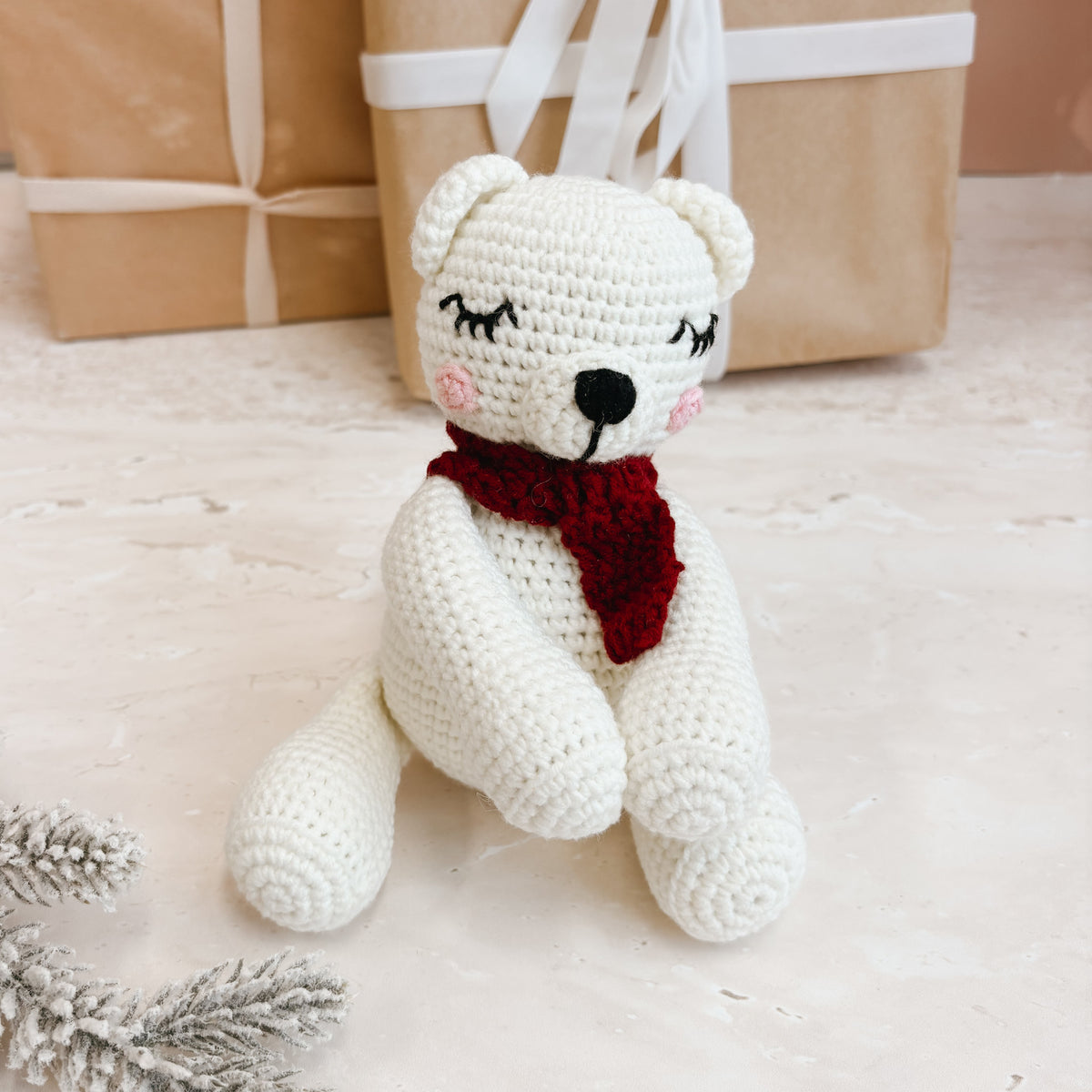 A crocheted white polar bear toy with a red scarf sits on a light surface, with wrapped gift boxes and silver pine branches in the background.