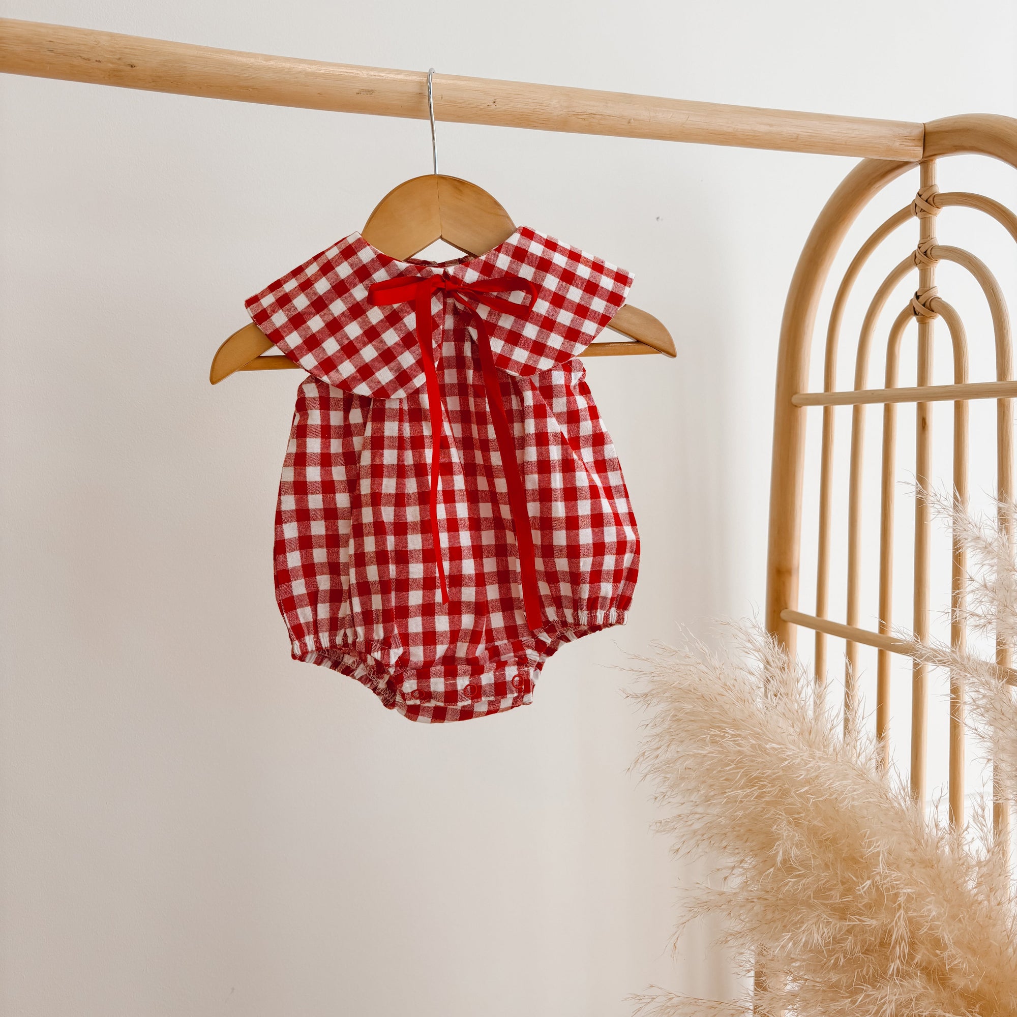 Red gingham baby romper with an oversized collar and red ribbon tie, hanging on a wooden hanger