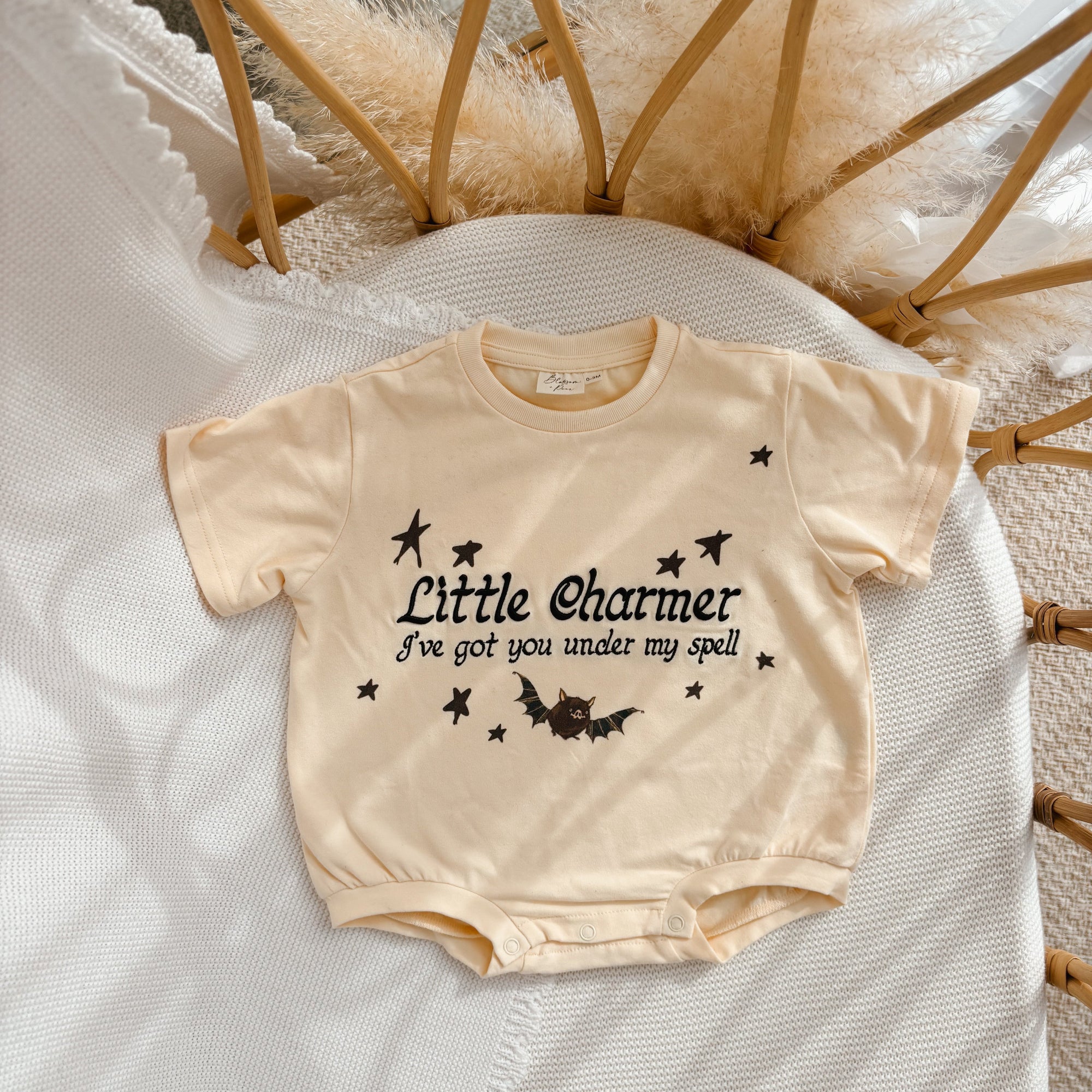 A cream-colored short-sleeve baby romper with a black graphic print reading 'Little Charmer I've got you under my spell' with stars and a bat, laid flat on a white textured blanket.