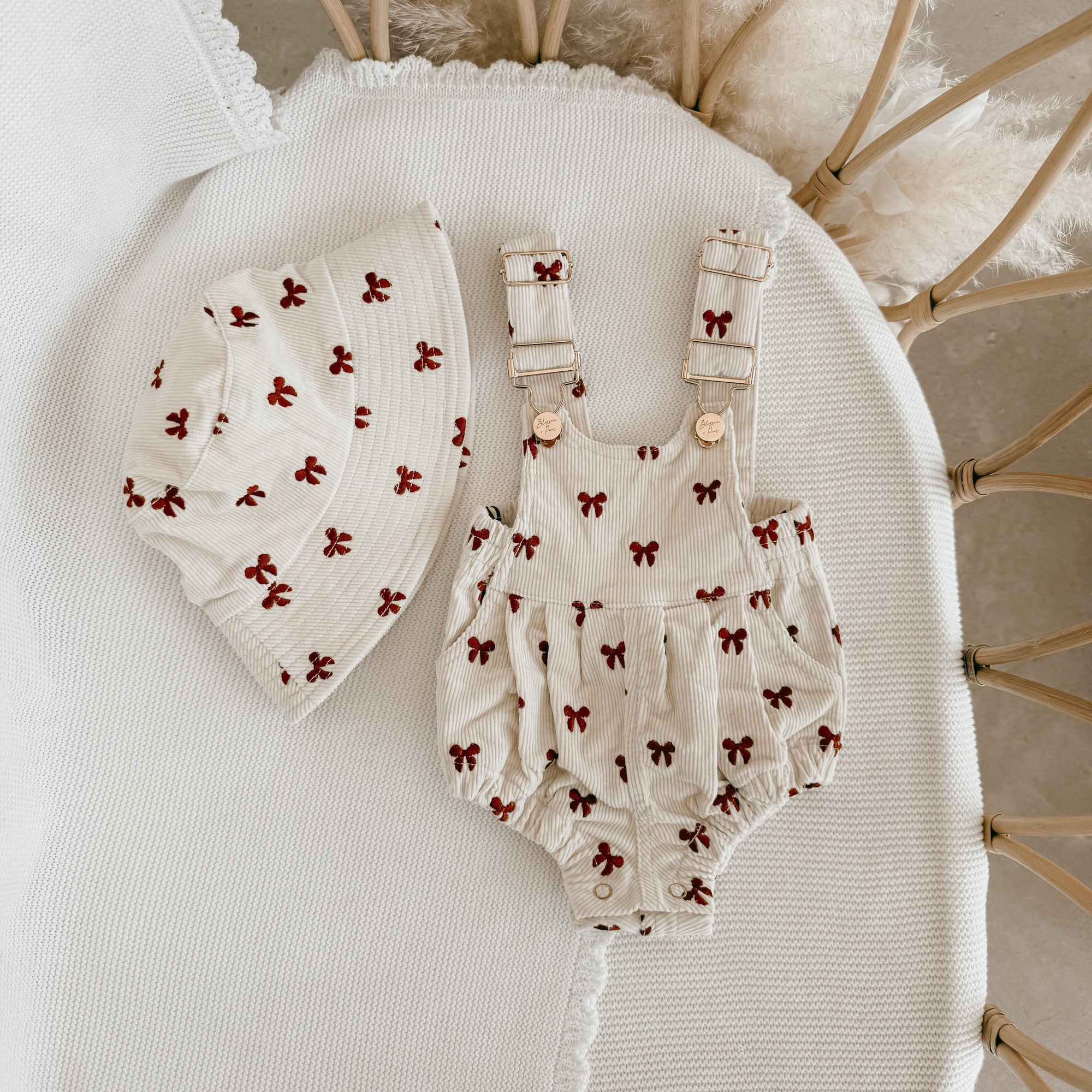 Red bow print corduroy baby overall romper with a matching bucket hat, flat lay in a bassinet