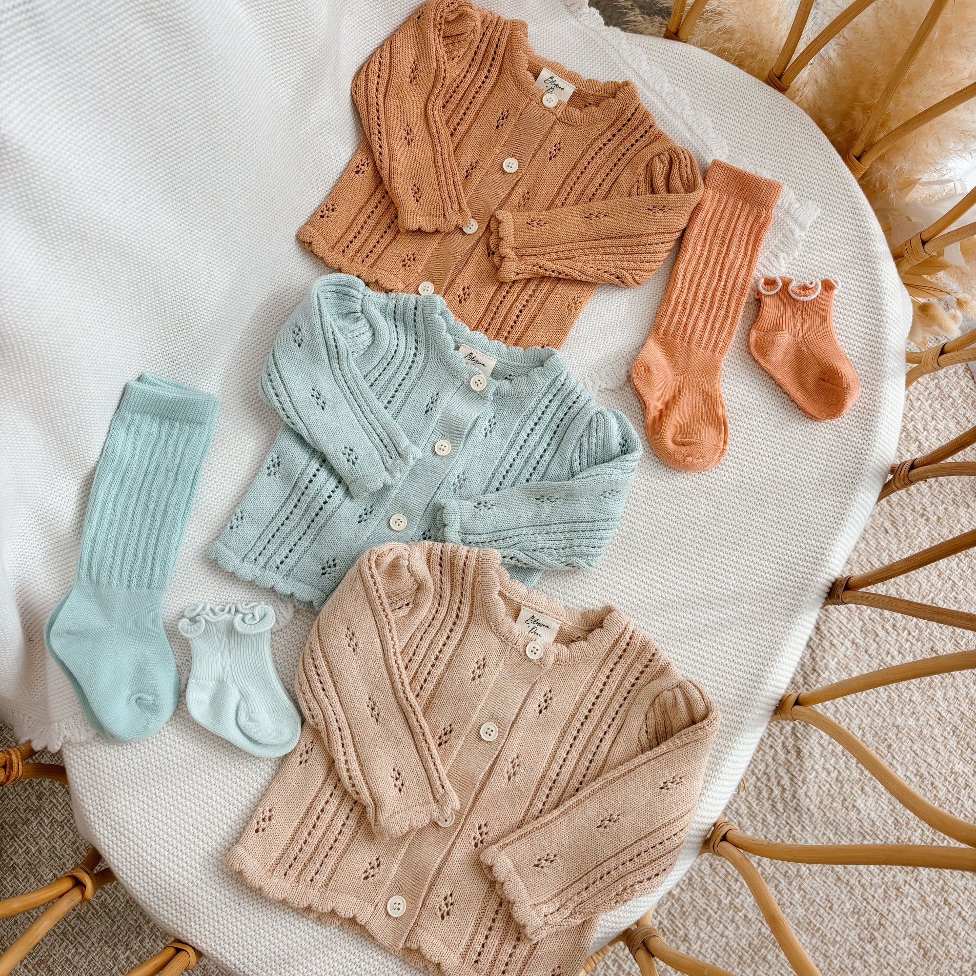 Three pointelle knit baby cardigans in tan, blue and oat with matching knee high socks, flat lay in a bassinet