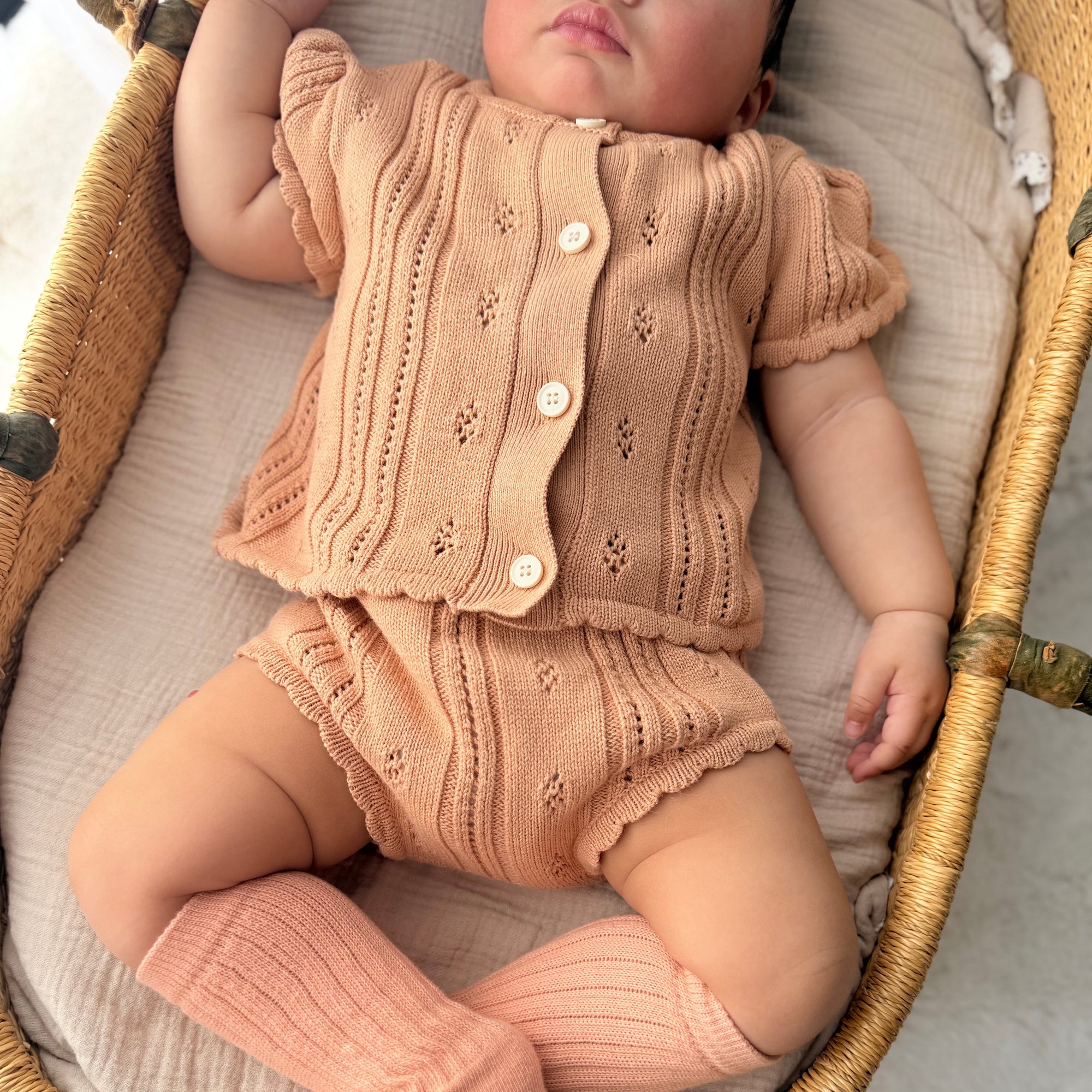 Baby in an apricot pointelle knit button top and bloomers with matching knee high socks in a bassinet