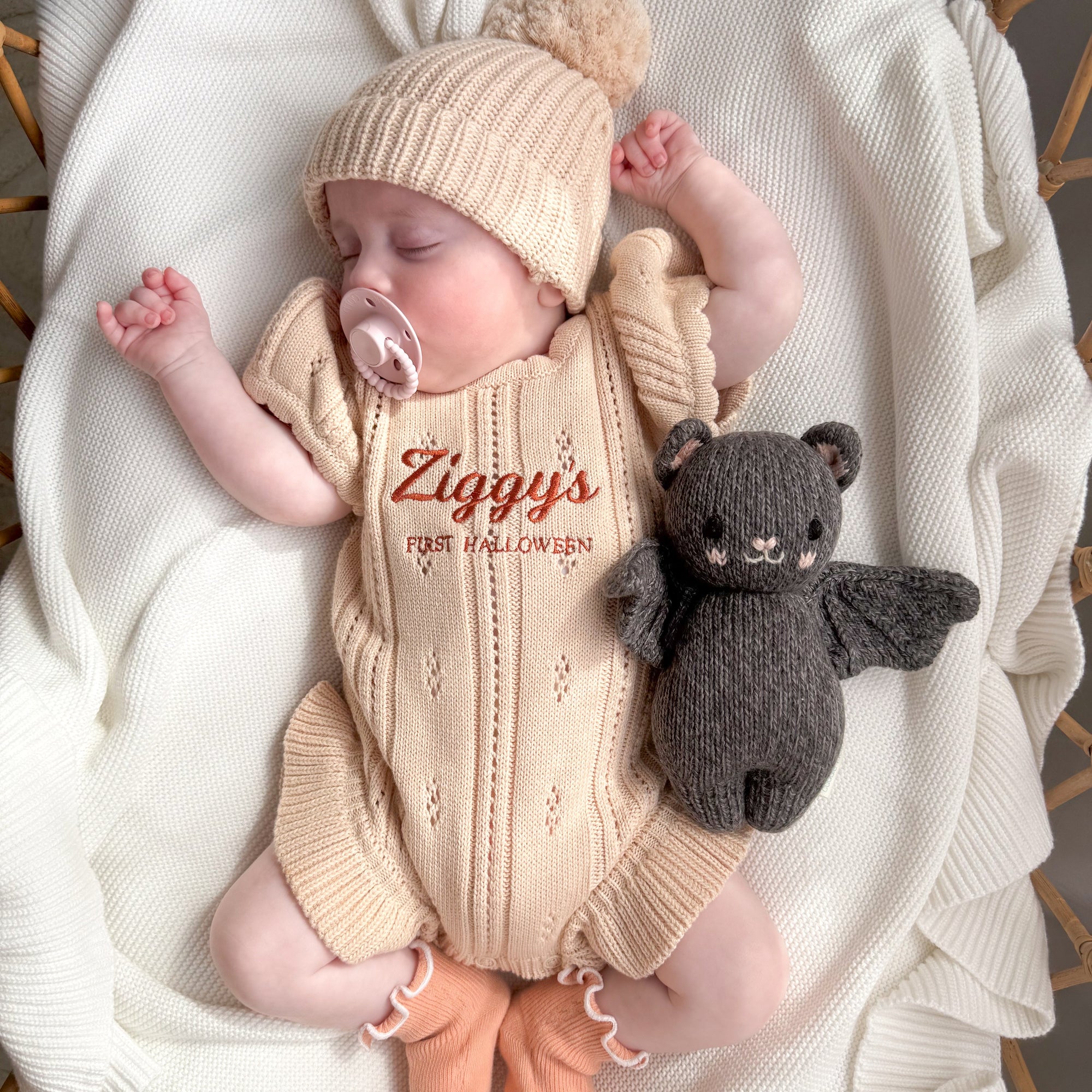 Sleeping baby in a personalised knit romper embroidered First Halloween, with a pom beanie and knitted bat toy