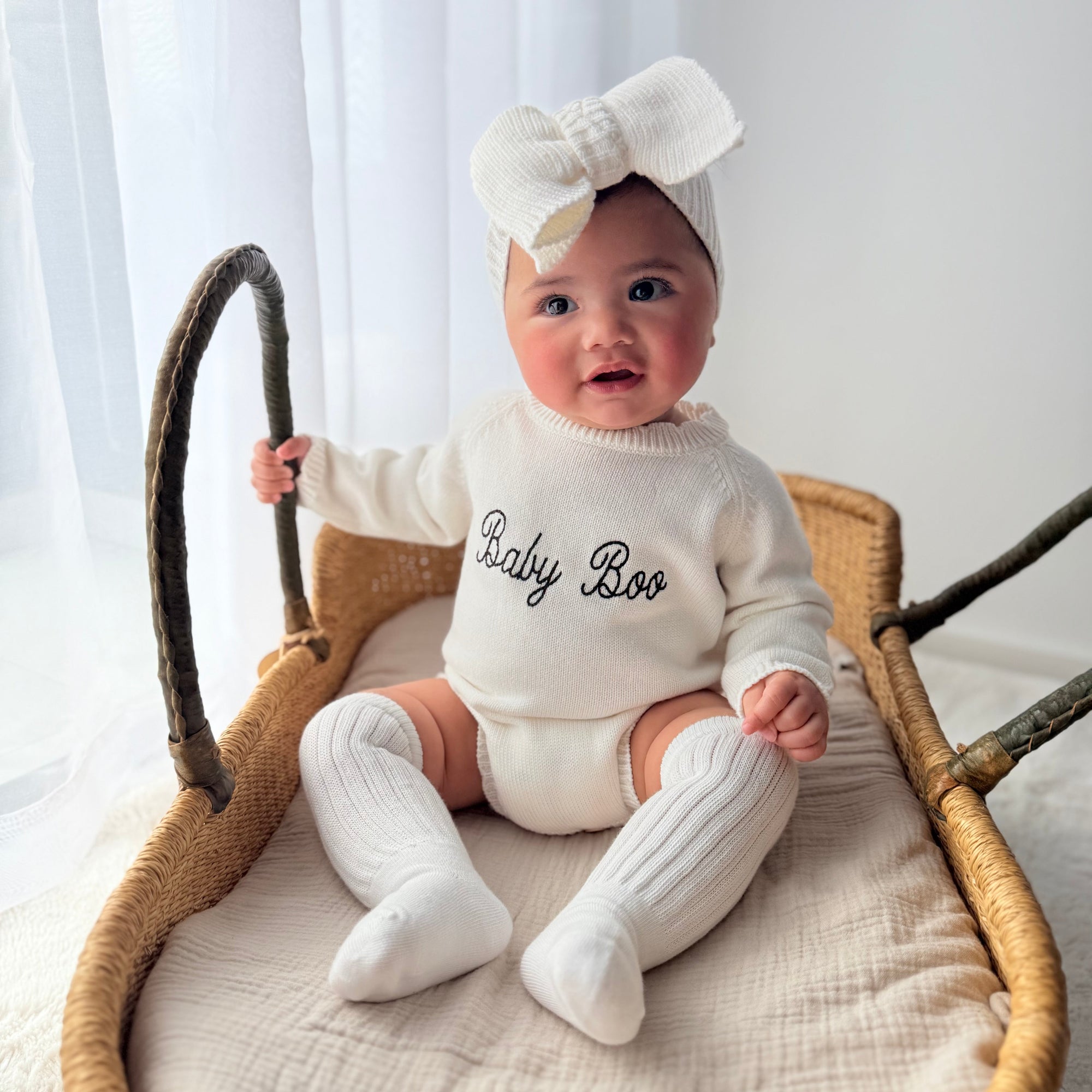Baby in a cream knit romper embroidered Baby Boo and a matching bow headband, sitting in a woven basket