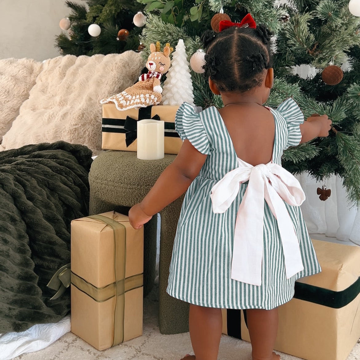 Back view of a toddler in a green stripe Christmas dress with a large white bow tie, decorating a tree