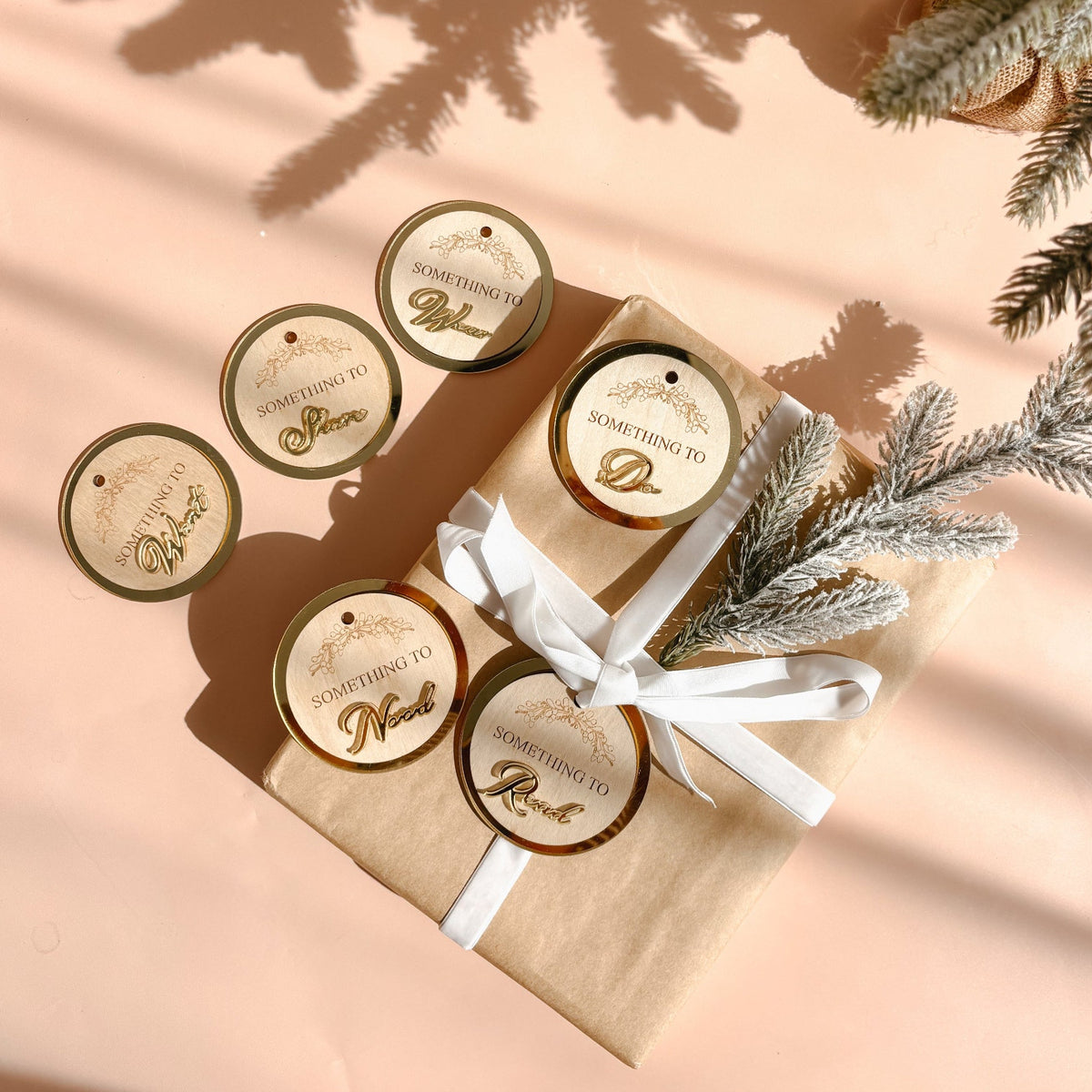 A styled flat-lay of round kraft paper gift tags with gold foil lettering, shown both loose and attached to a brown paper gift box tied with a white ribbon. The scene includes decorative pine branches and strong diagonal shadows on a peach-colored surface.
