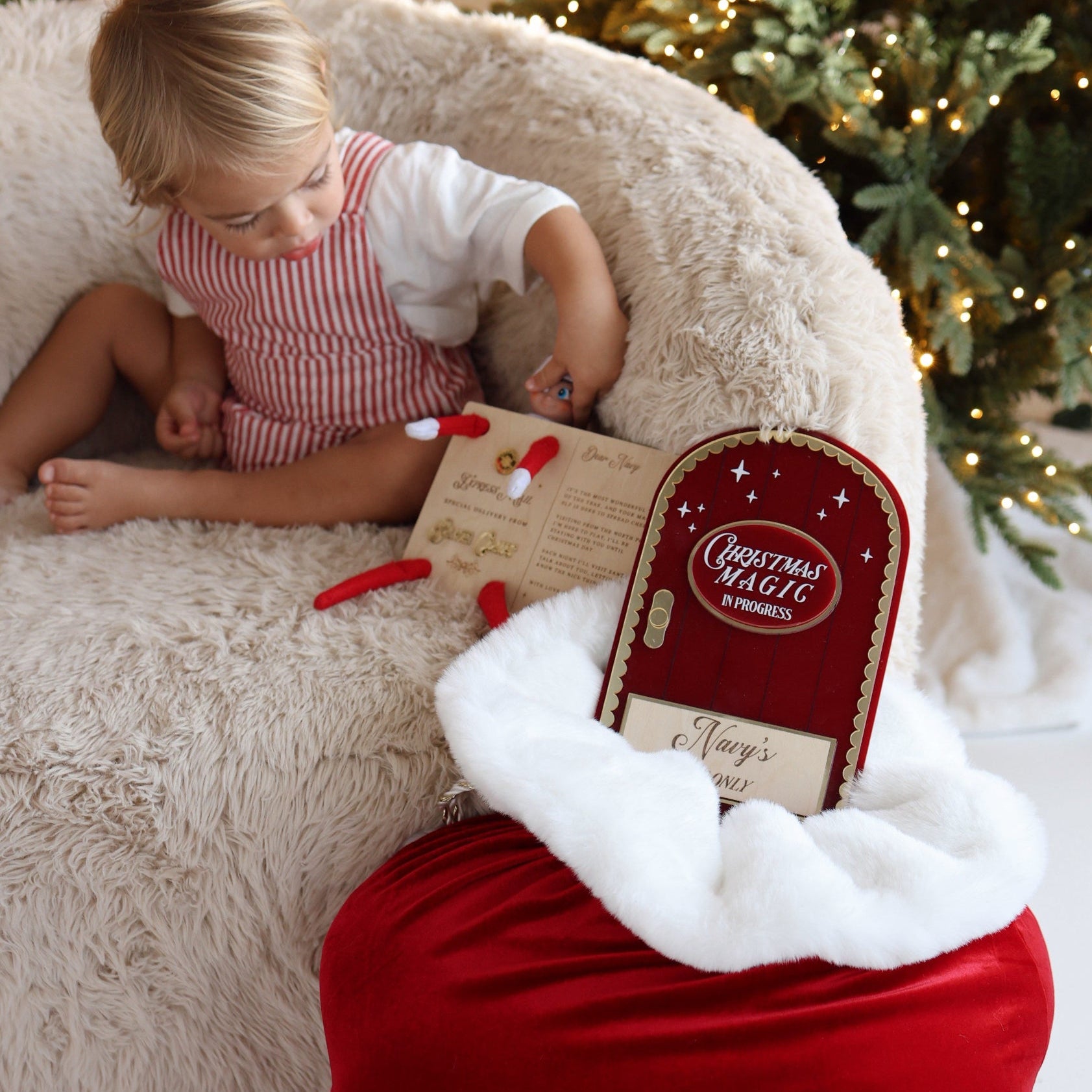 A child sits on a fluffy cream chair beside a red Christmas sack with white trim and gold script reading 'Navy'. A small gift box labeled 'Christmas Magic' is placed in the sack, with a decorated Christmas tree in the background.