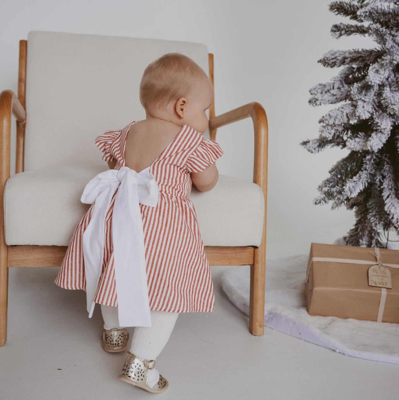 Back view of a baby in a red stripe Christmas dress with a large white bow tie, beside a flocked tree