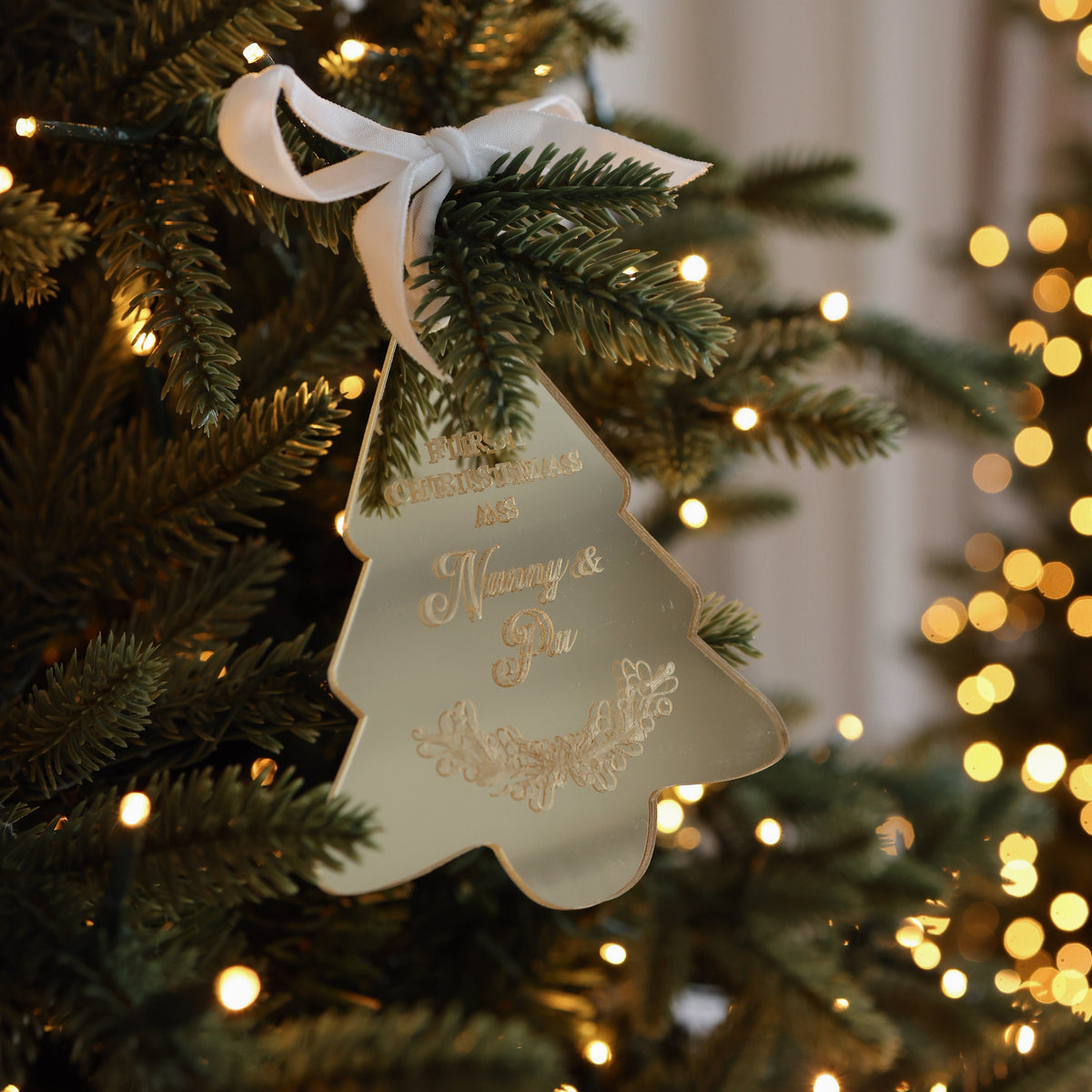 Mirrored acrylic tree ornament engraved 'First Christmas as Nanny &amp; Pa', tied with a white ribbon to a lit Christmas tree