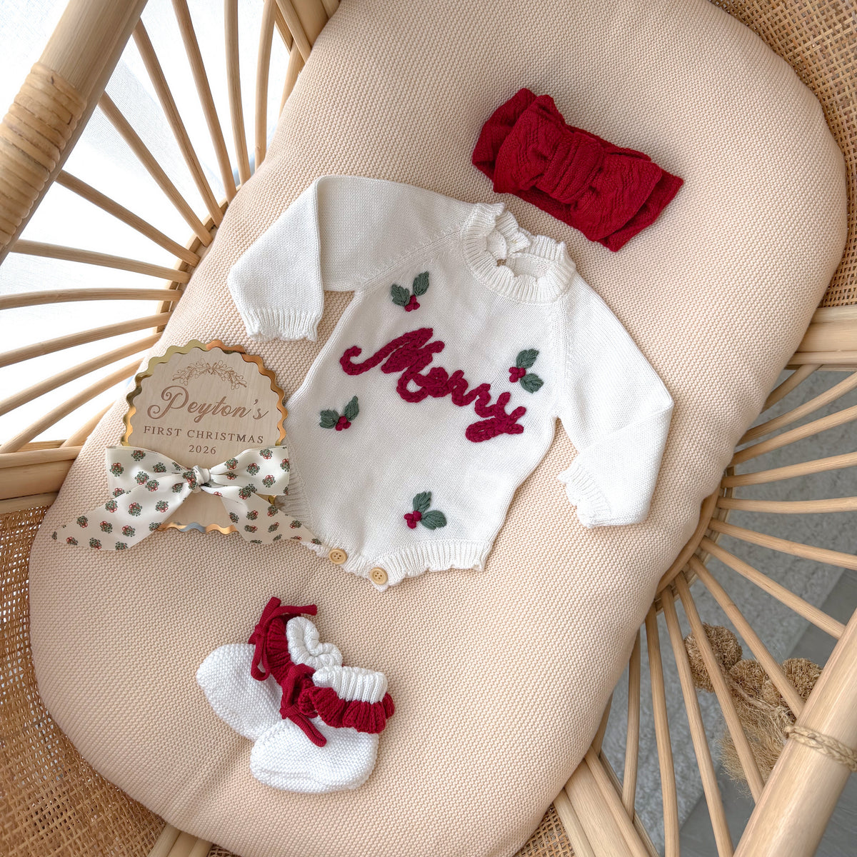 Cream Merry knit romper flat lay with a red knot headband, knit booties, a holly print bow and a personalised Peyton's First Christmas 2026 keepsake disc