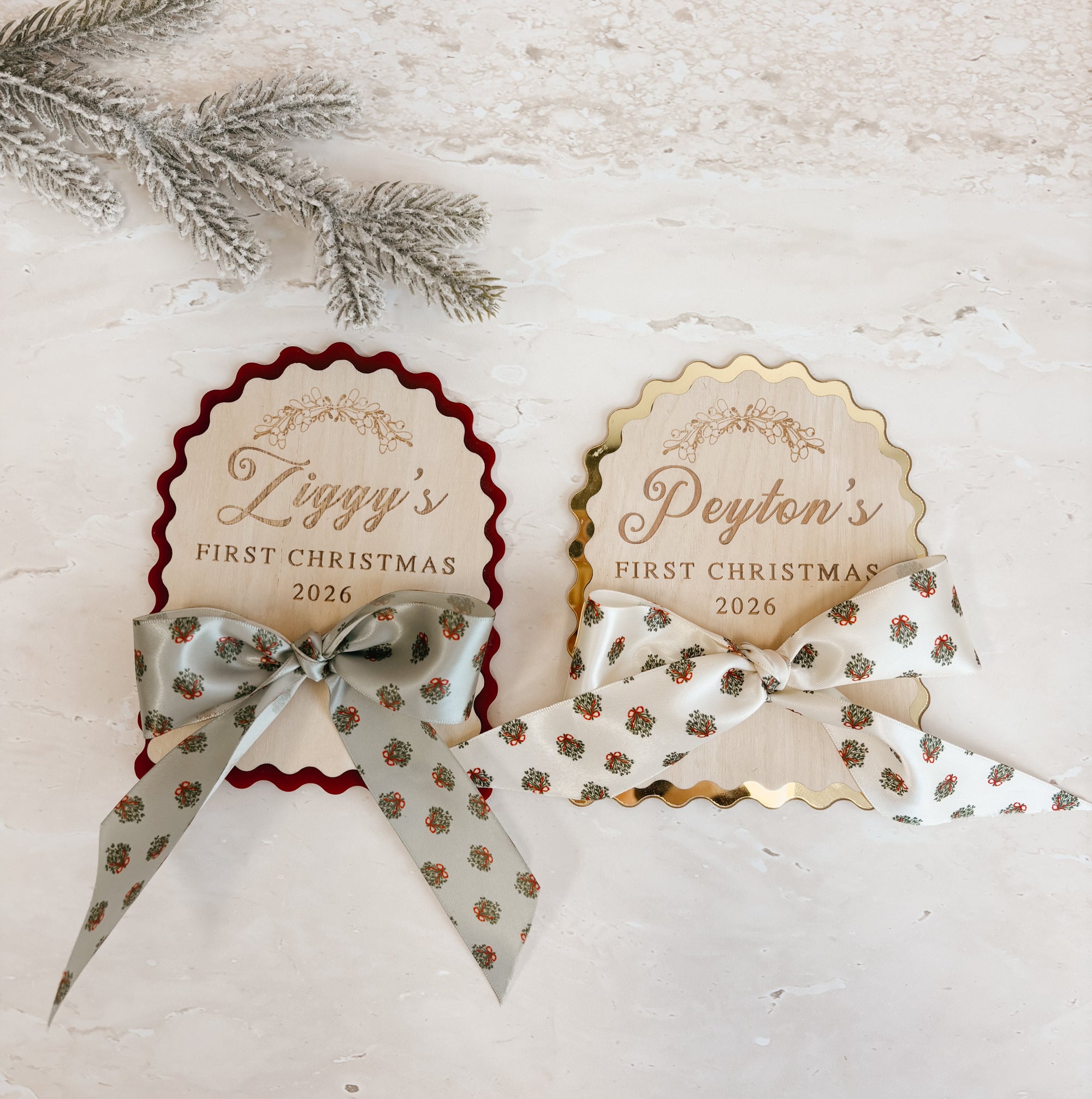 Two personalized Christmas favor tags with ribbon bows, displayed on a textured light beige surface with silver pine branches. One tag has a dark red scalloped border and the other has a gold scalloped border, both with beige backgrounds and elegant script text.