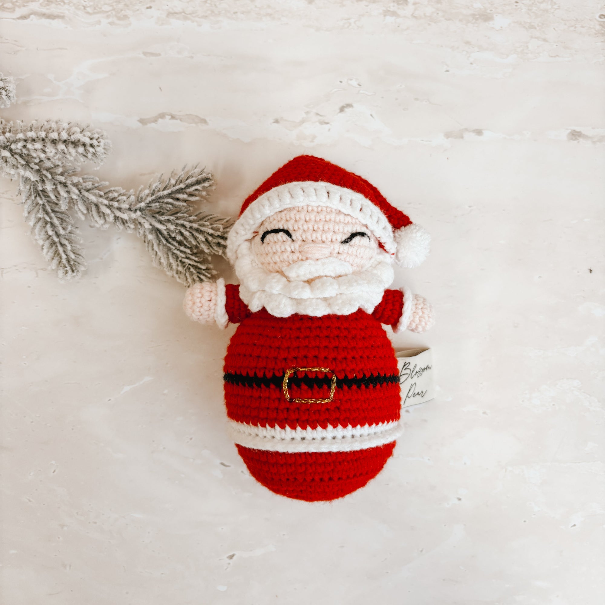 A crocheted Santa Claus figure with a red and white body, white beard, and red hat, lying on a textured off-white surface next to a small silver pine branch.