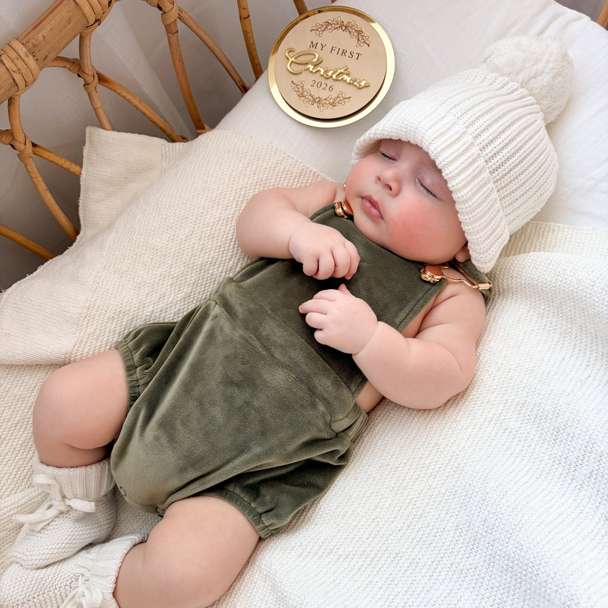 Sleeping baby in an olive velvet romper and white pom beanie beside a gold My First Christmas 2026 keepsake disc